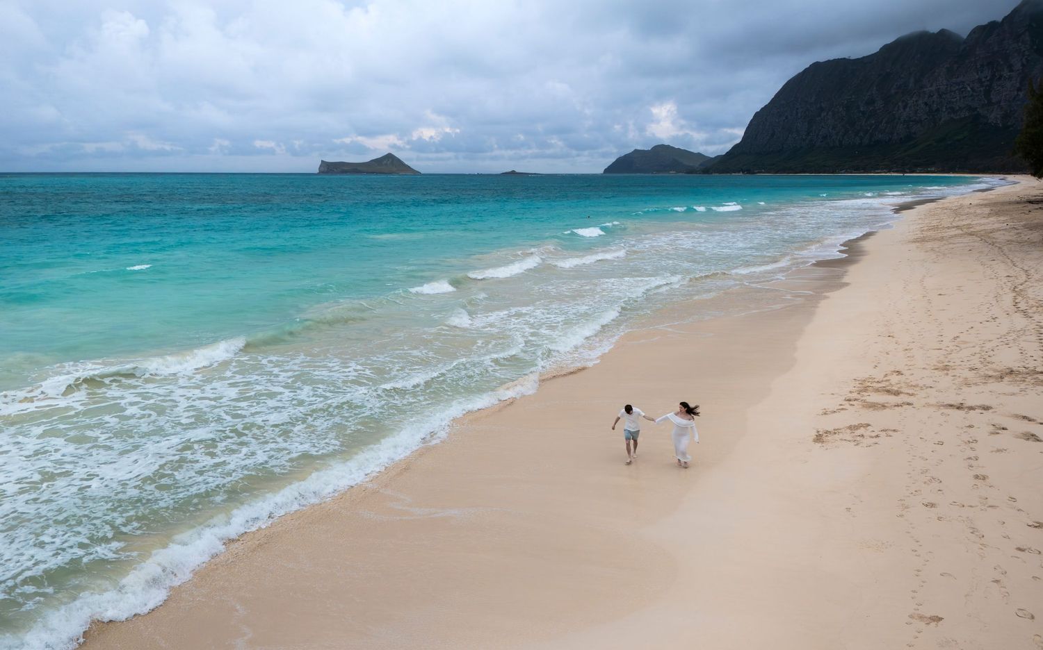 Couple walking hand in hand on a pristine sandy beach with turquoise waters and dramatic mountains in Hawaii.