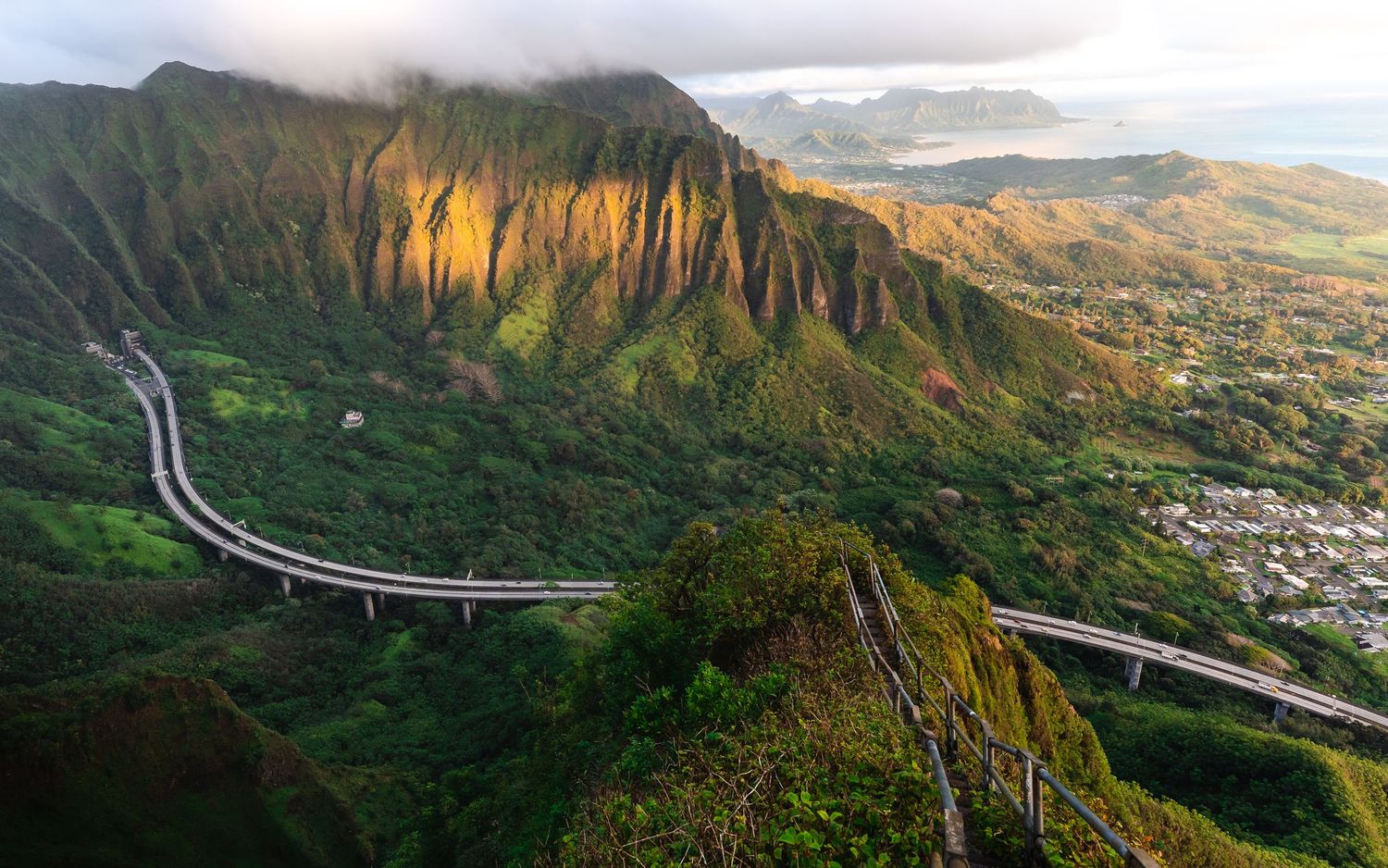 Dramatic aerial view of mountain cliffs and winding roads along the Hawaiian coastline at sunset.