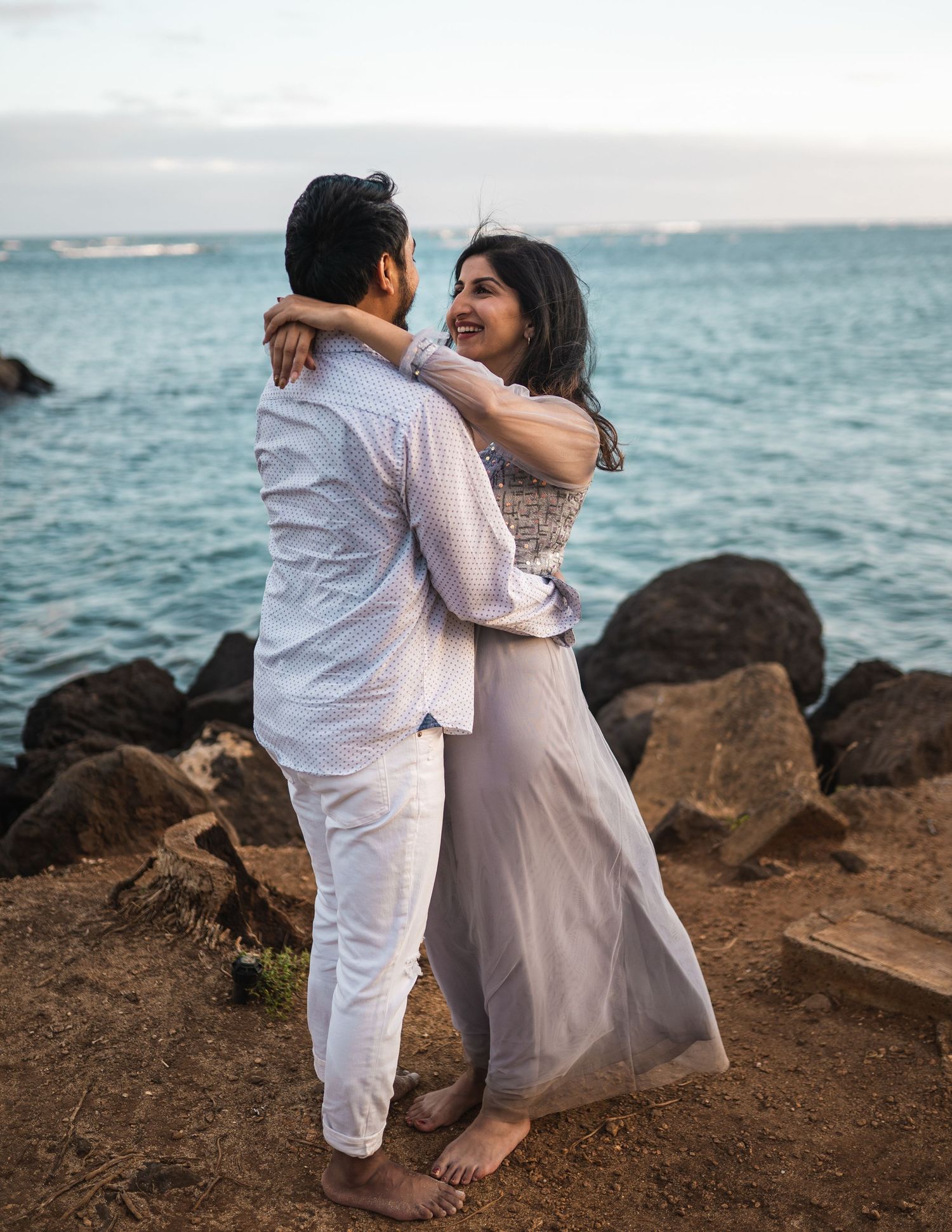 Couple embracing on rocky coastline with turquoise ocean waves in the background.
