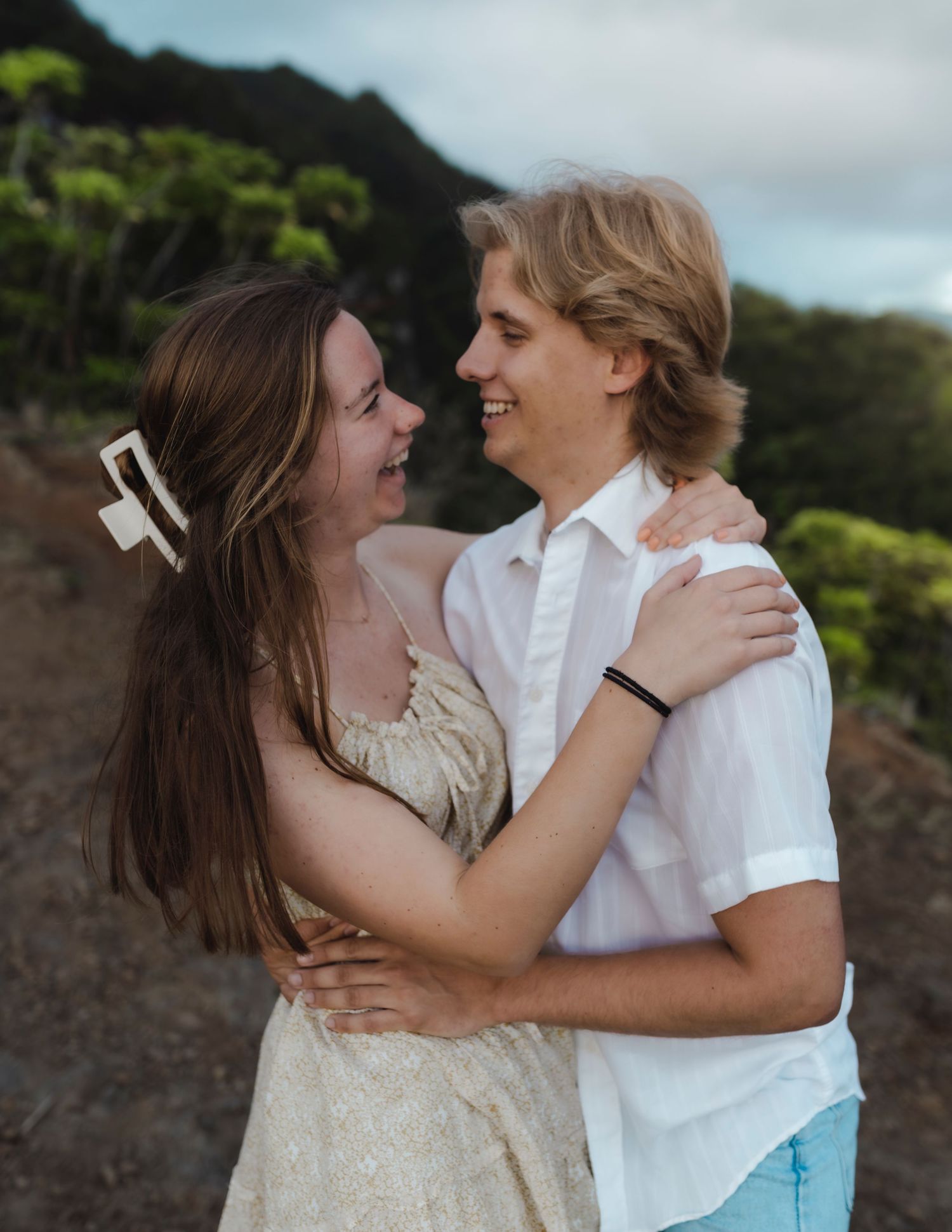A romantic couple embraces and smiles at each other against a mountainous backdrop at sunset.