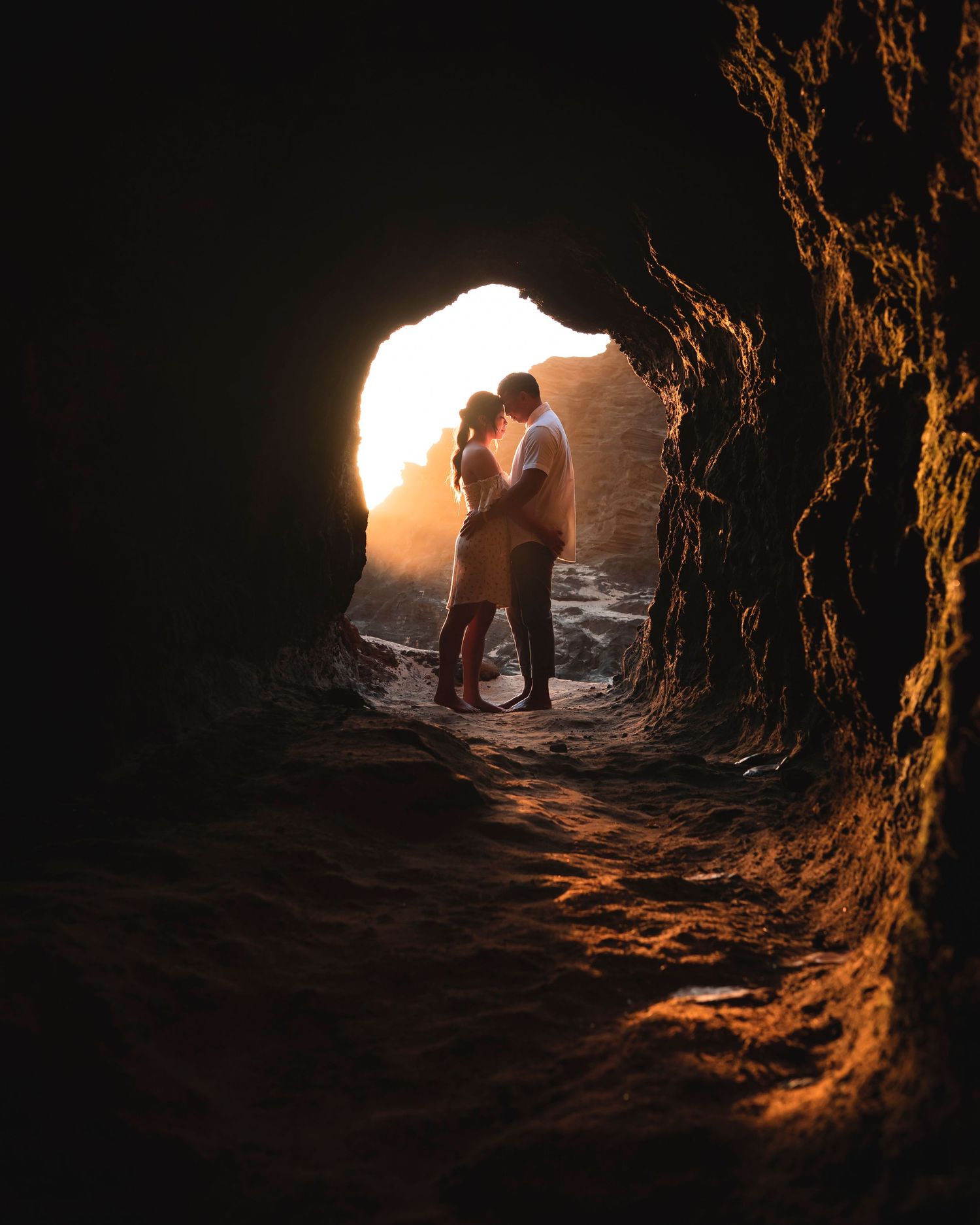 Couple embracing romantically at the entrance of a dark sea cave, backlit by warm golden sunset light.