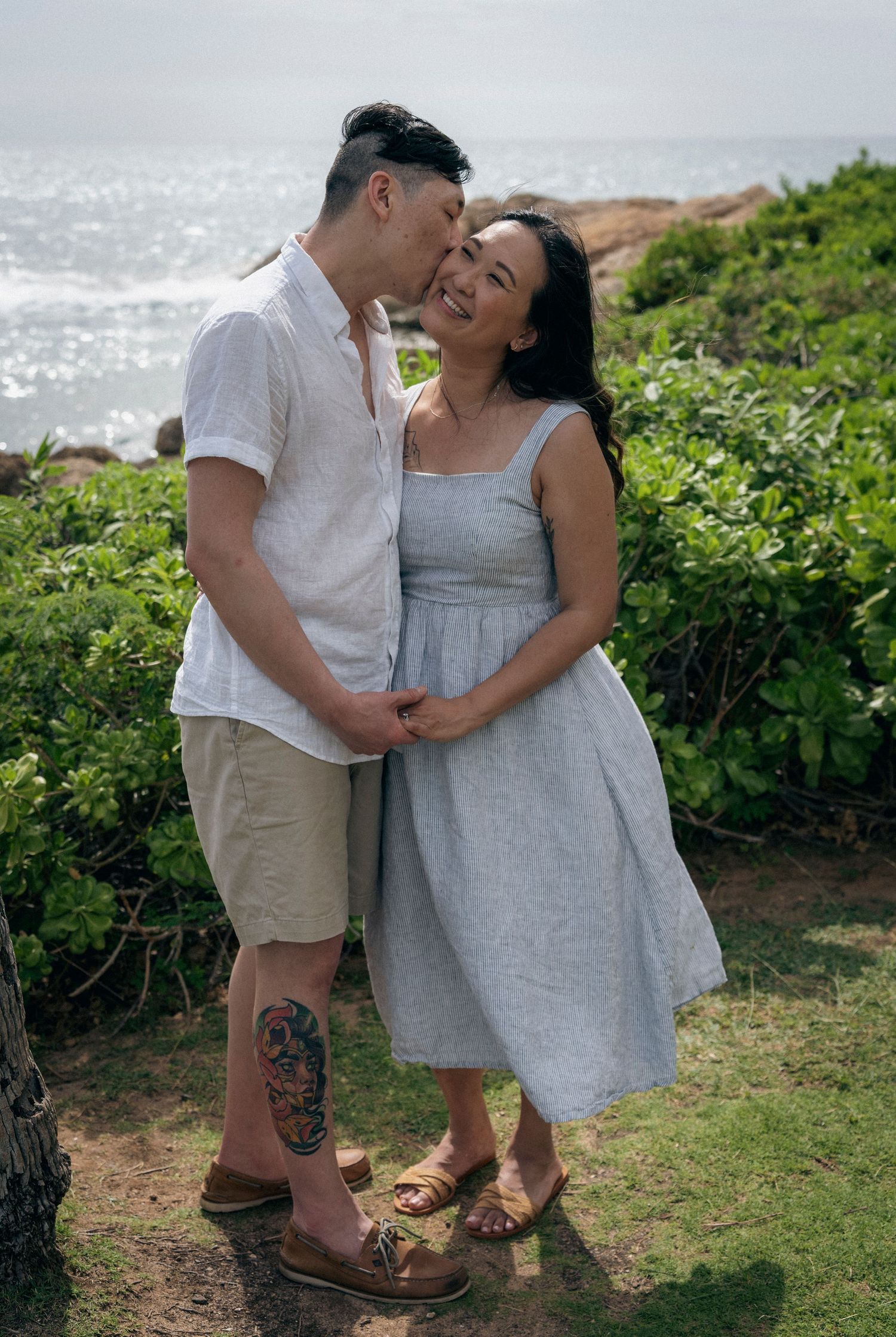 Romantic couple embracing on coastal cliff with ocean views and green foliage.