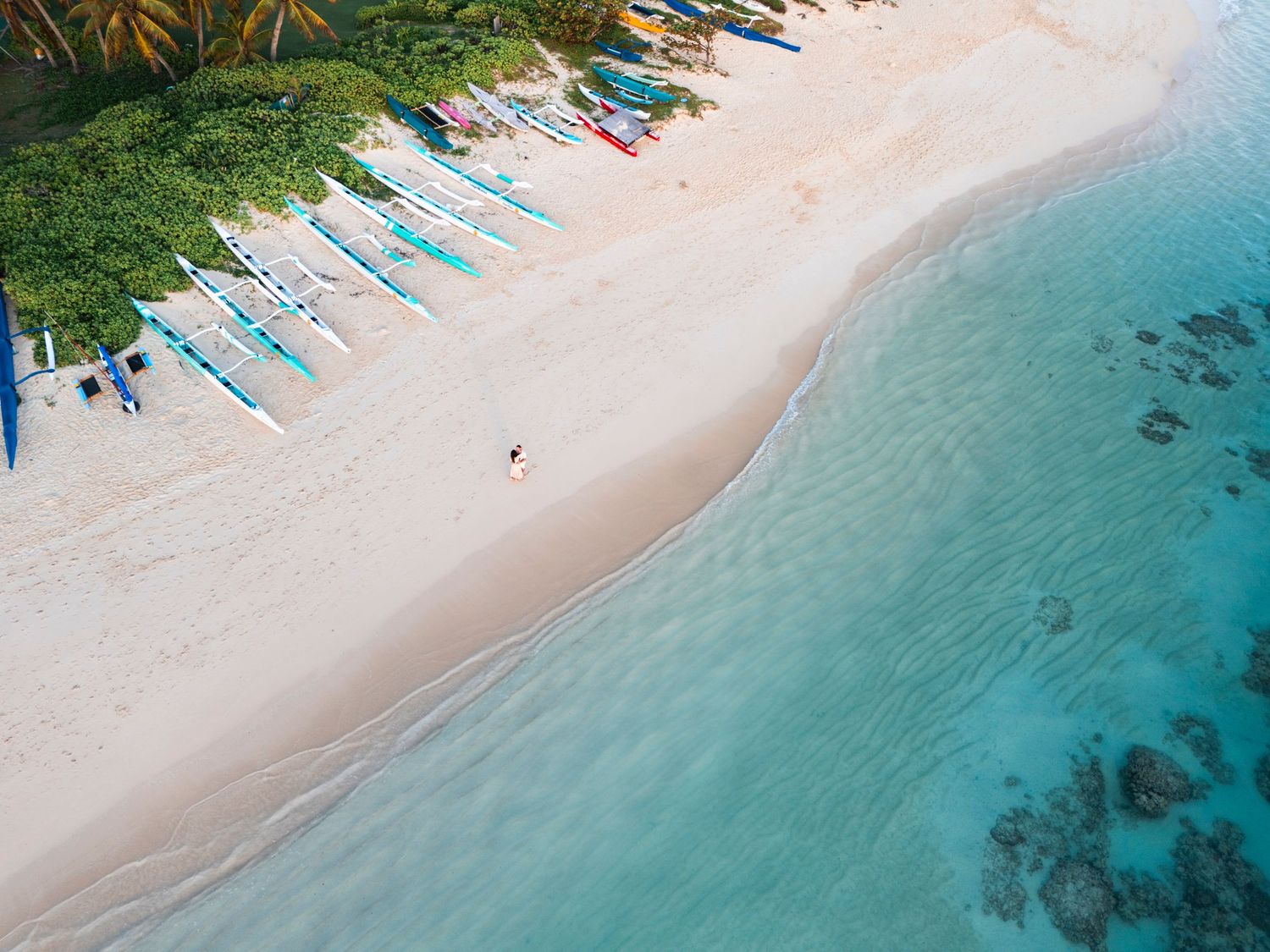 Aerial view of a tropical white sand beach with colorful outrigger boats lined up along the shore and turquoise water.
