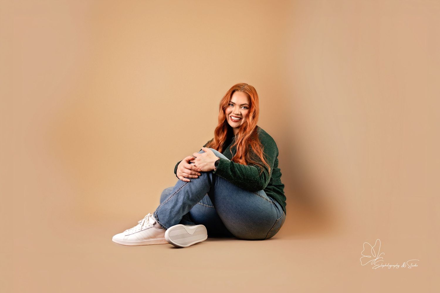 A figure in a green sweater and jeans sits casually on a wooden floor near a window. Branding photography in Williston, North Dakota.