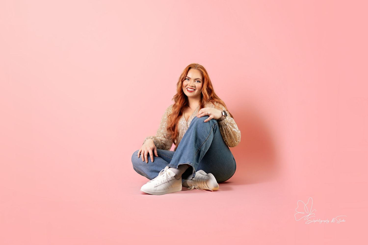 A person sitting on a pink backdrop wearing jeans and white sneakers. Branding photography in Williston, North Dakota.