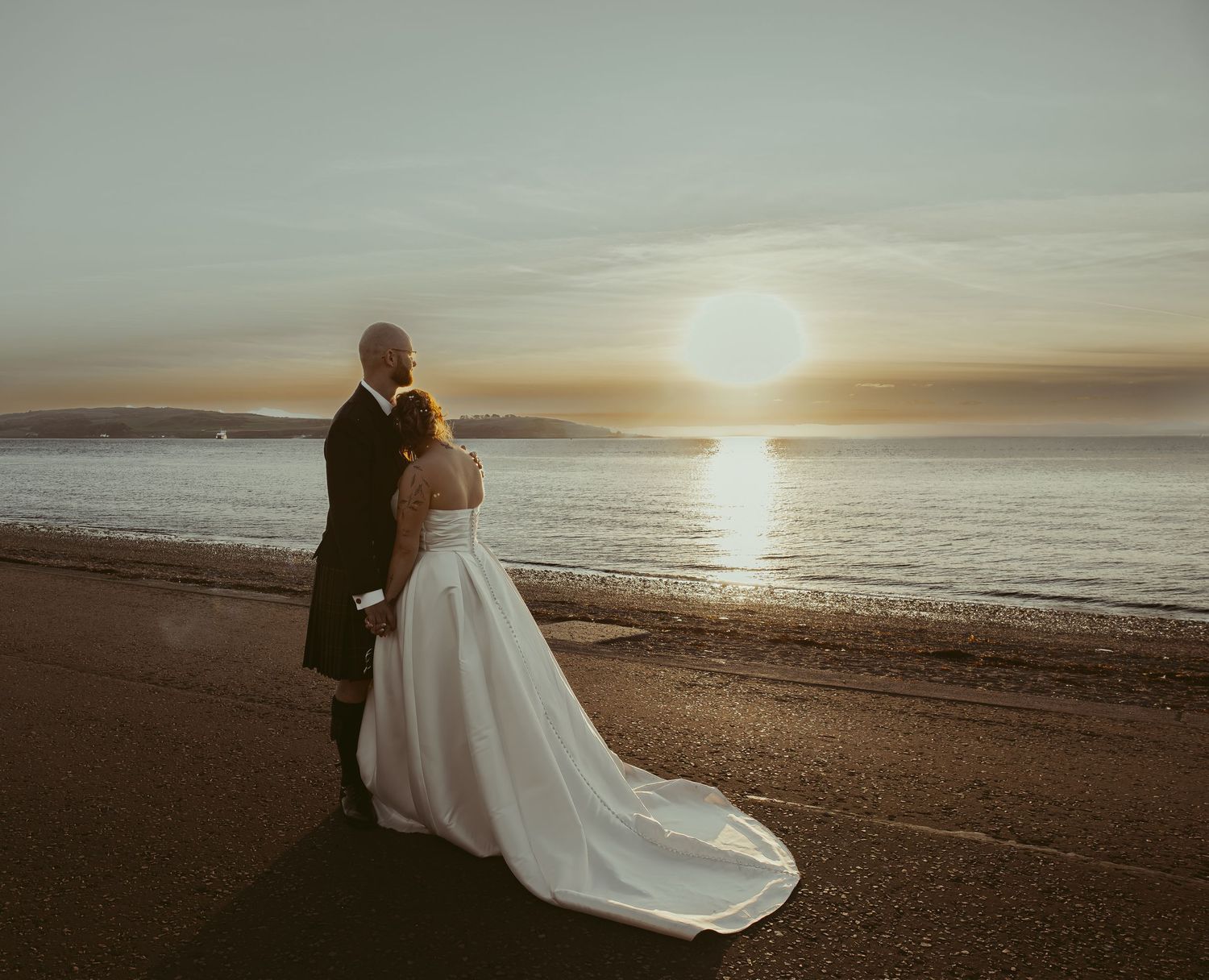 Bride and groom embrace at sunset on a beach, with golden light reflecting on calm ocean waters.