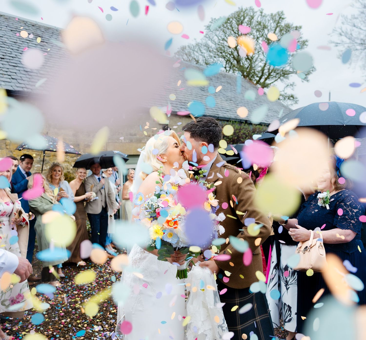 Bride and groom kissing amid colorful confetti shower surrounded by wedding guests holding umbrellas outdoors.