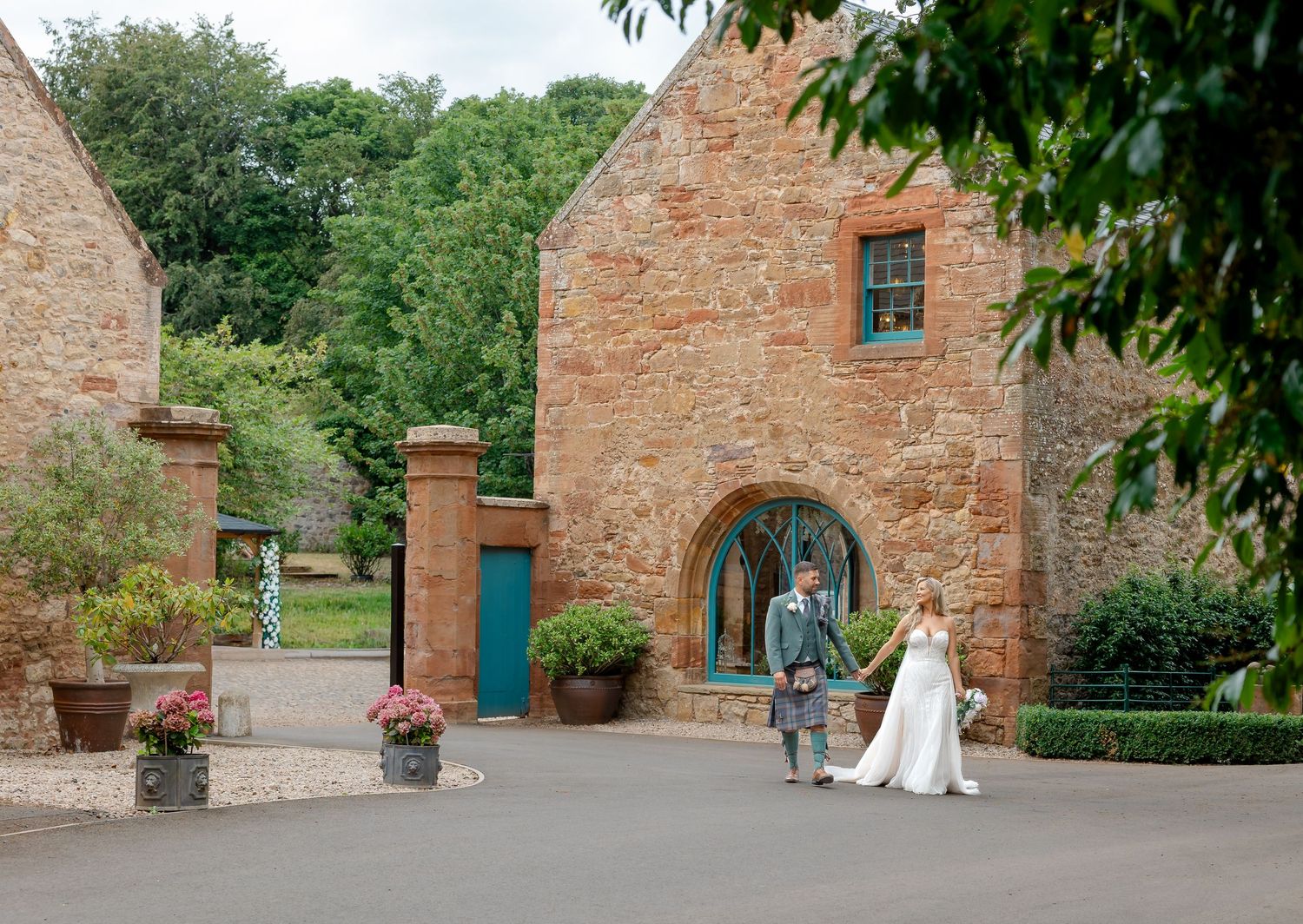 Bride and groom walking hand in hand outside a rustic red sandstone wedding venue surrounded by lush greenery.