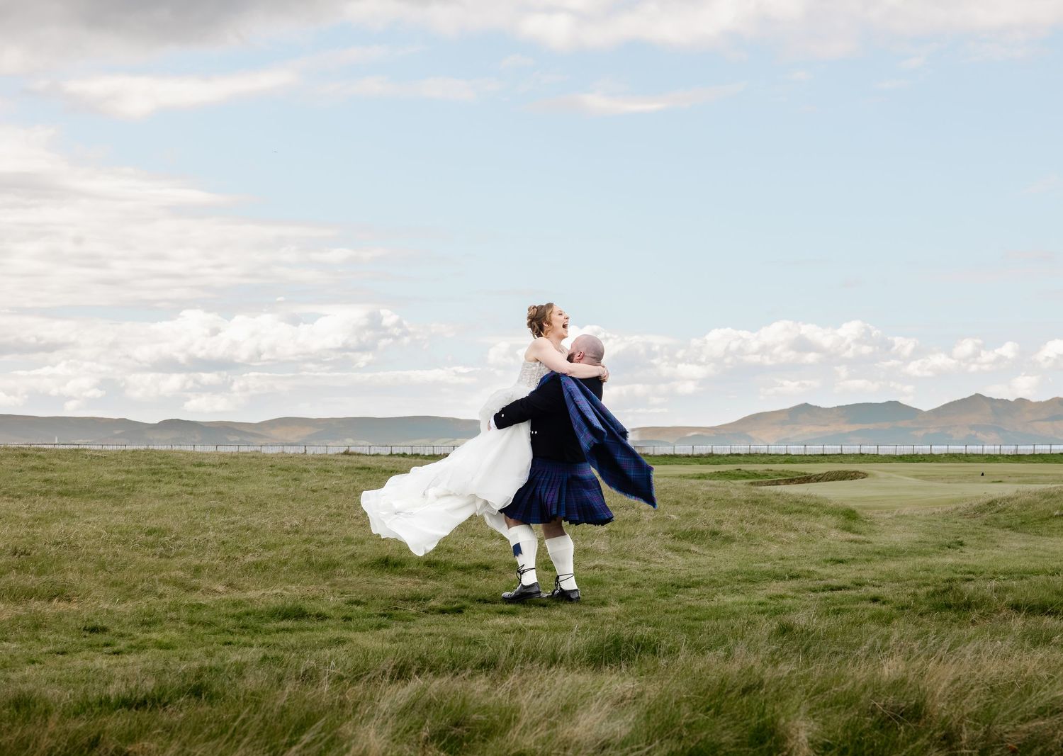 Groom in navy kilt lifts bride in white dress in a scenic green field with hills and cloudy sky.