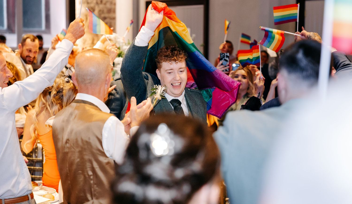 Joyful wedding celebration with guests waving rainbow Pride flags around a smiling groom in a festive venue.