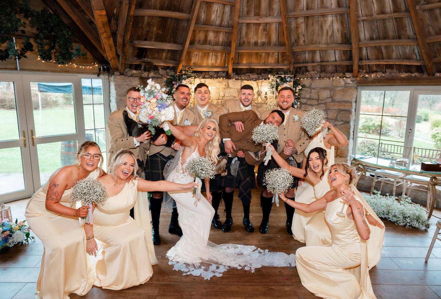 Wedding party celebrating indoors, bridesmaids in ivory gowns and groomsmen in kilts cheering with bouquets in rustic venue.