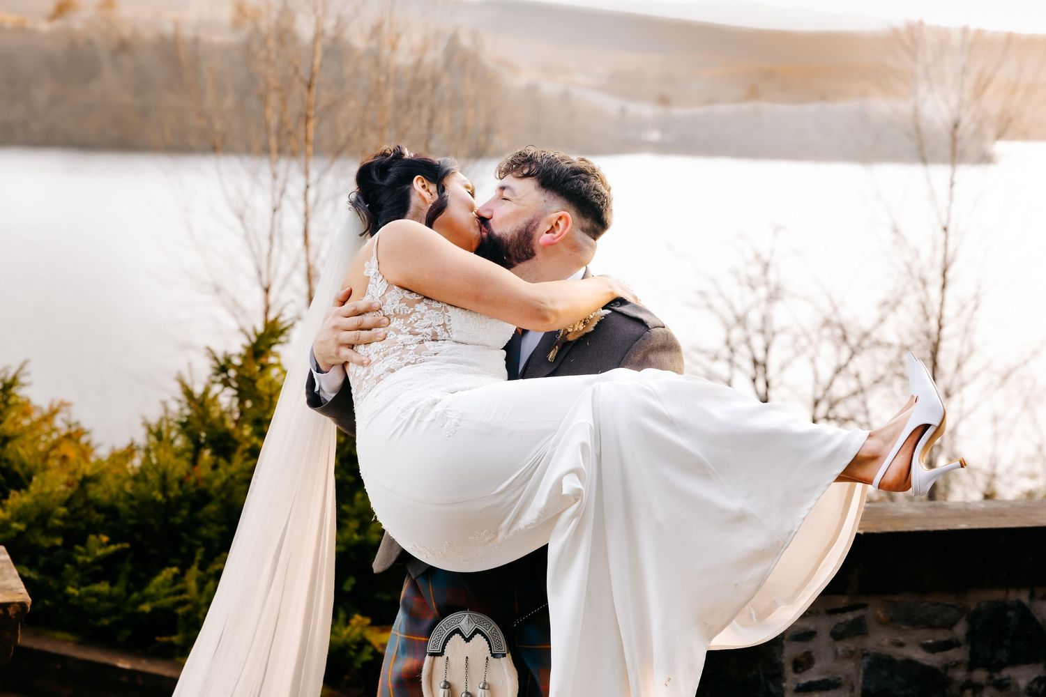 A couple embraces in a romantic pose on a snowy winter day with evergreen trees in the background.