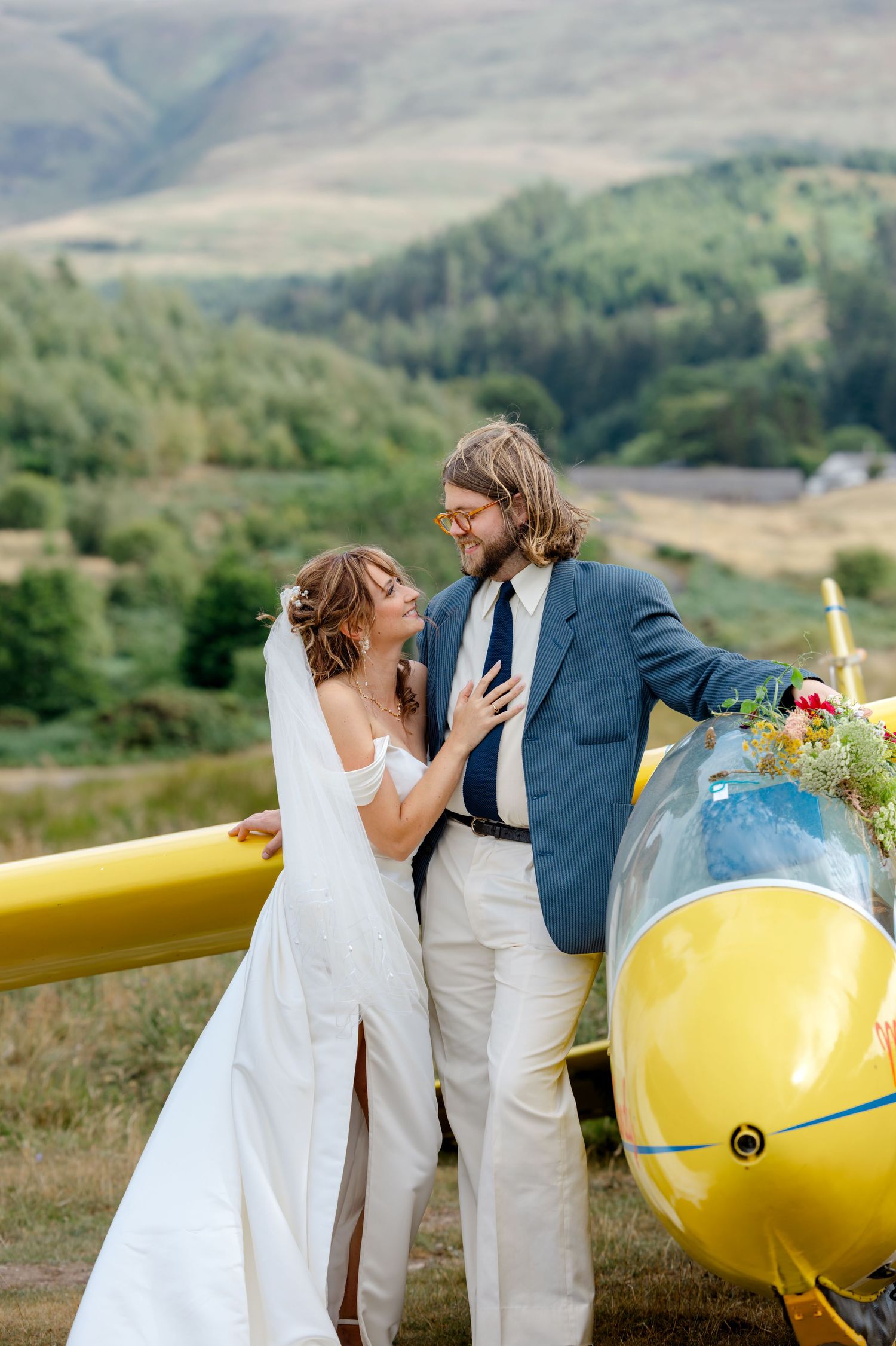 Bride and groom embrace beside a yellow glider plane with scenic green hills in the background at their outdoor wedding.