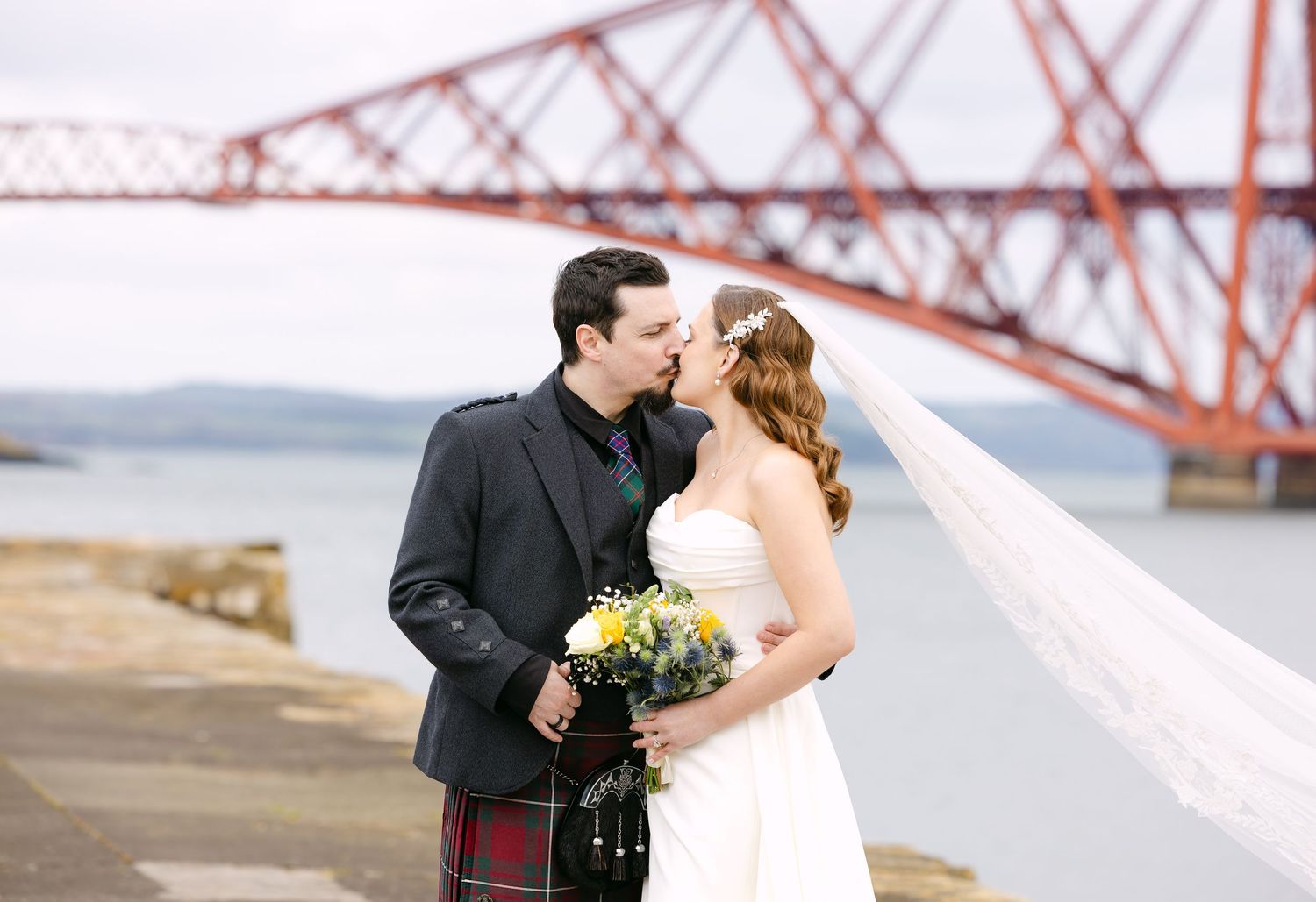 Bride and groom kissing near the Forth Rail Bridge in Scotland, groom wearing traditional tartan kilt.