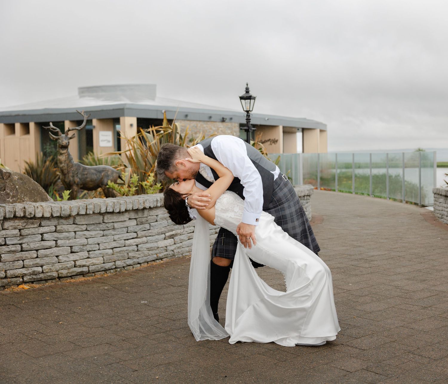 Bride and groom share a romantic dip kiss outdoors on their wedding day, with a misty venue backdrop.