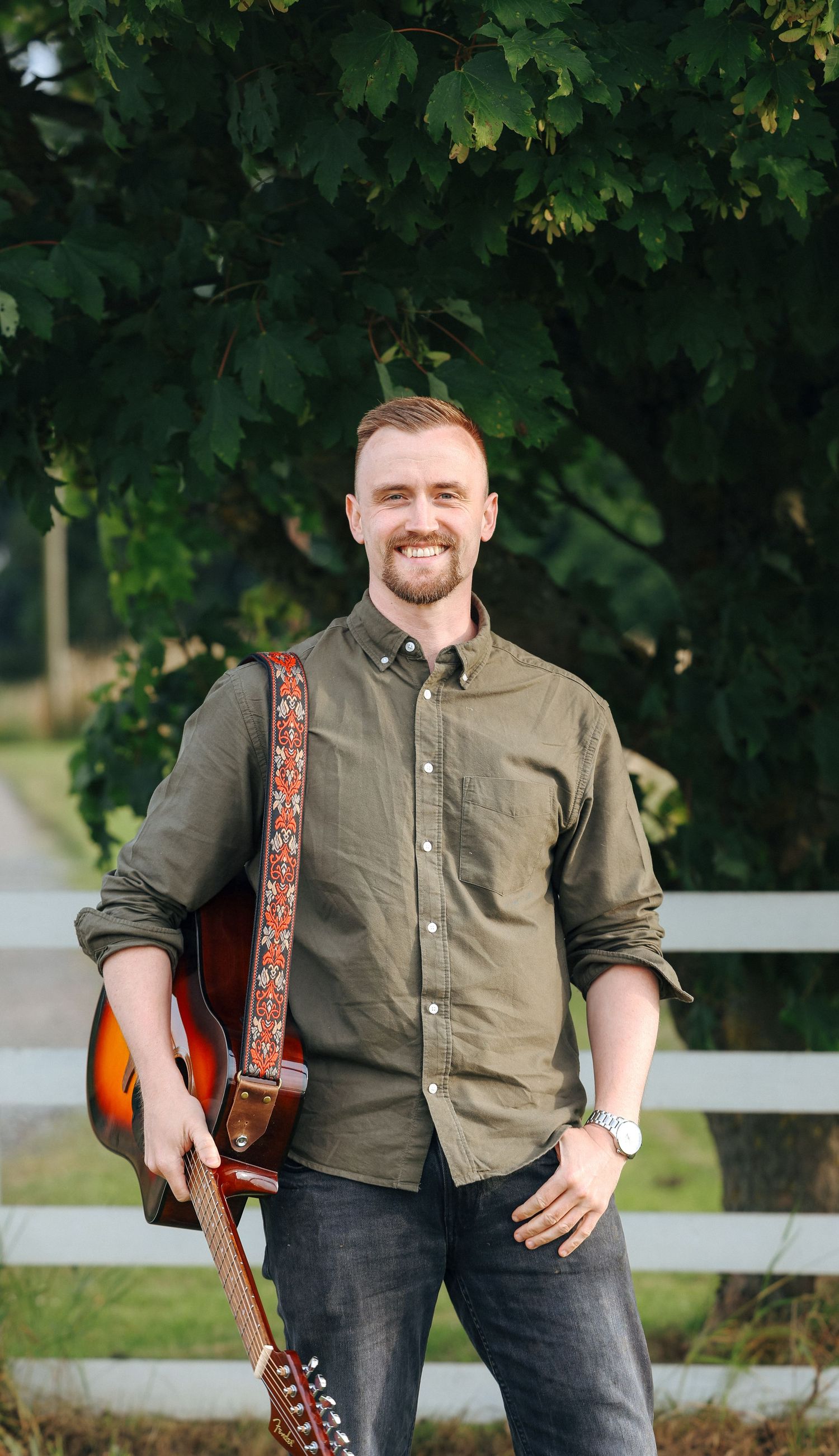 Smiling man with beard holding acoustic guitar with decorative strap, standing outdoors near white fence and green trees.