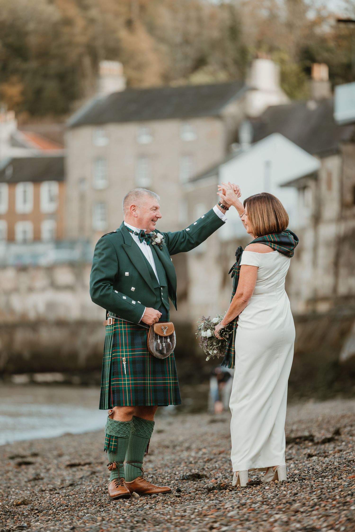 A couple in Scottish wedding attire sharing a playful moment on a rocky beach with stone buildings in the background.