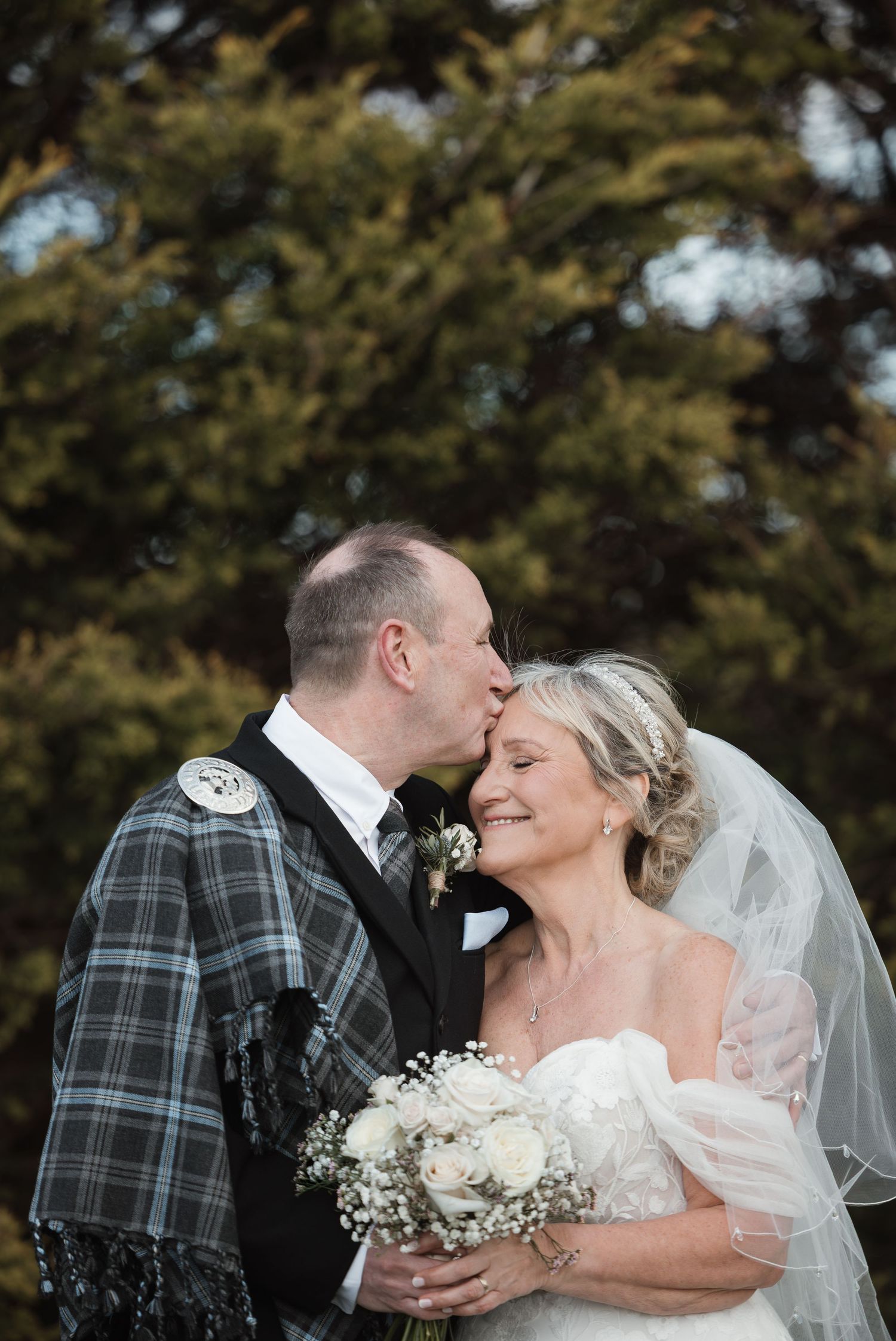 Groom in Scottish tartan kilt kisses bride wearing veil and holding white bouquet outdoors.