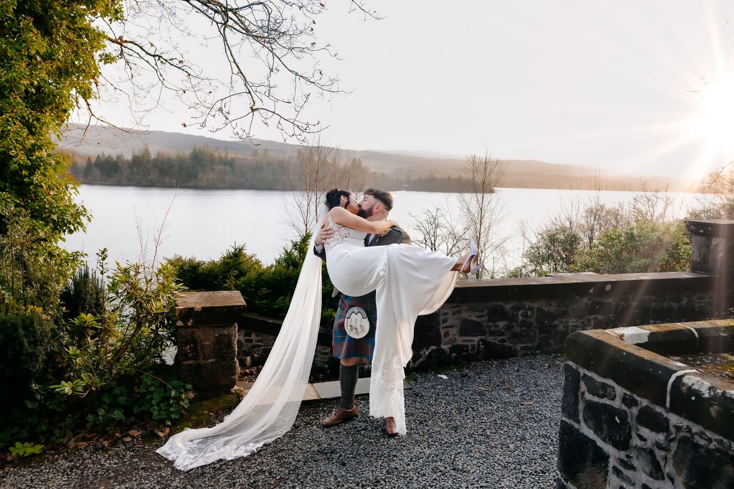 Groom lifting bride in white gown by lakeside at sunset, with scenic water and trees in background.