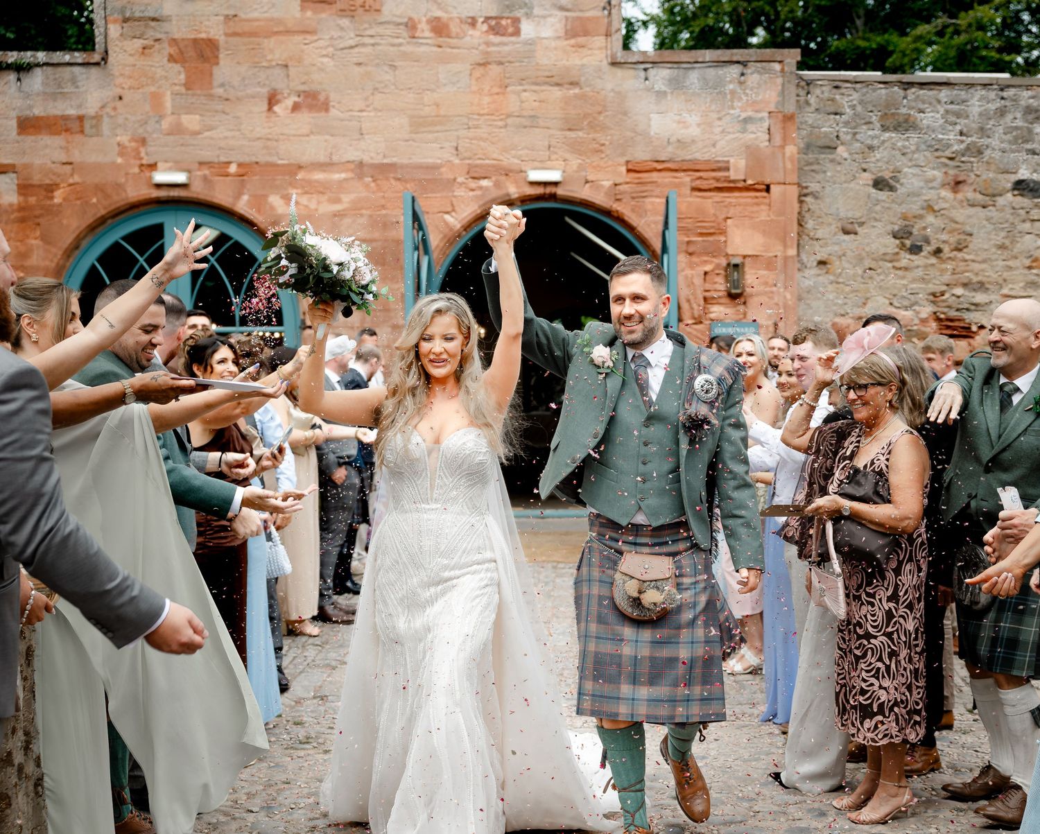 Bride and groom in kilt exit wedding ceremony through cheering guests at a rustic Scottish venue.