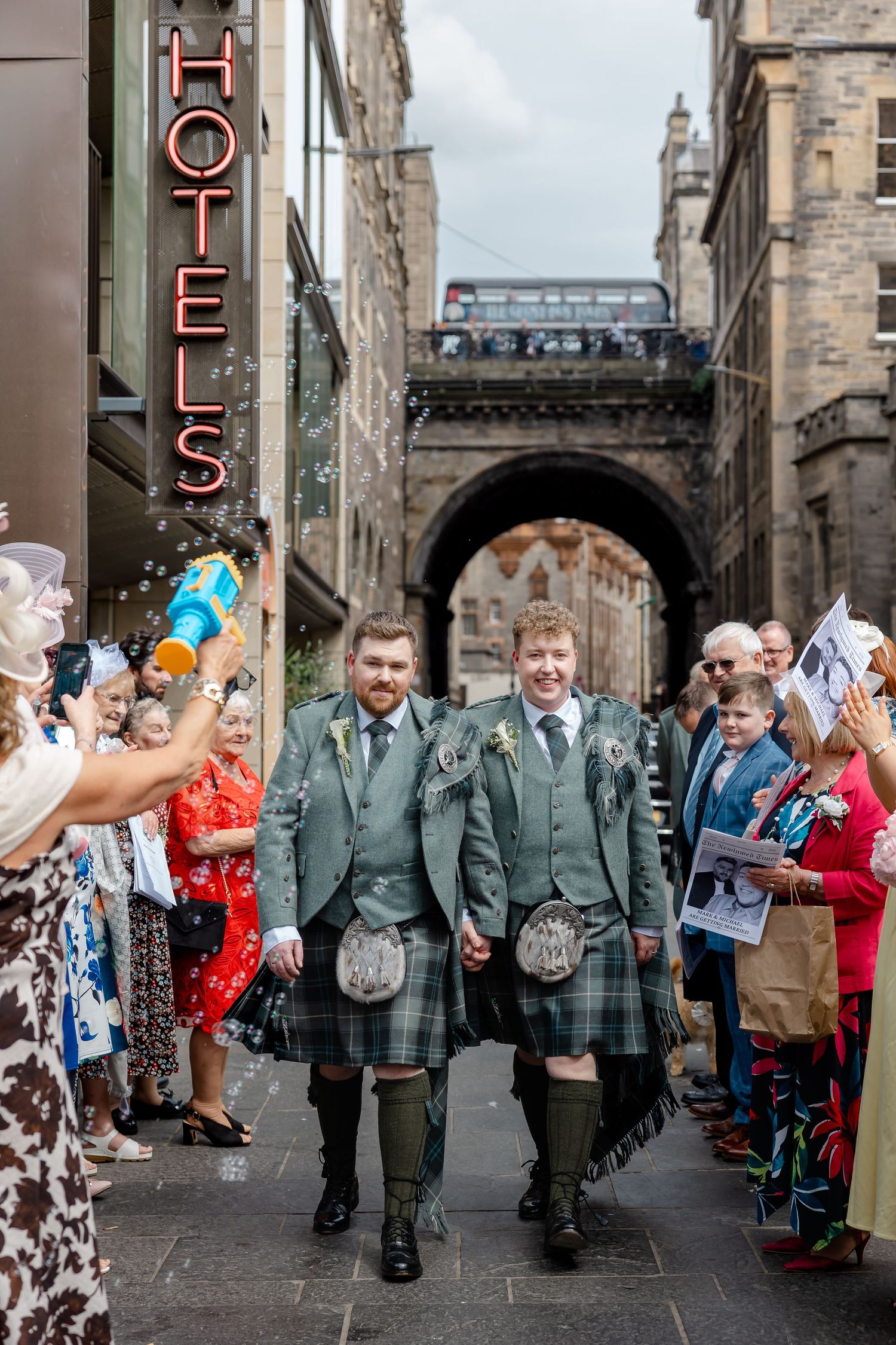 Two grooms in green Scottish kilts walk hand-in-hand through Edinburgh streets, celebrated by cheering wedding guests.
