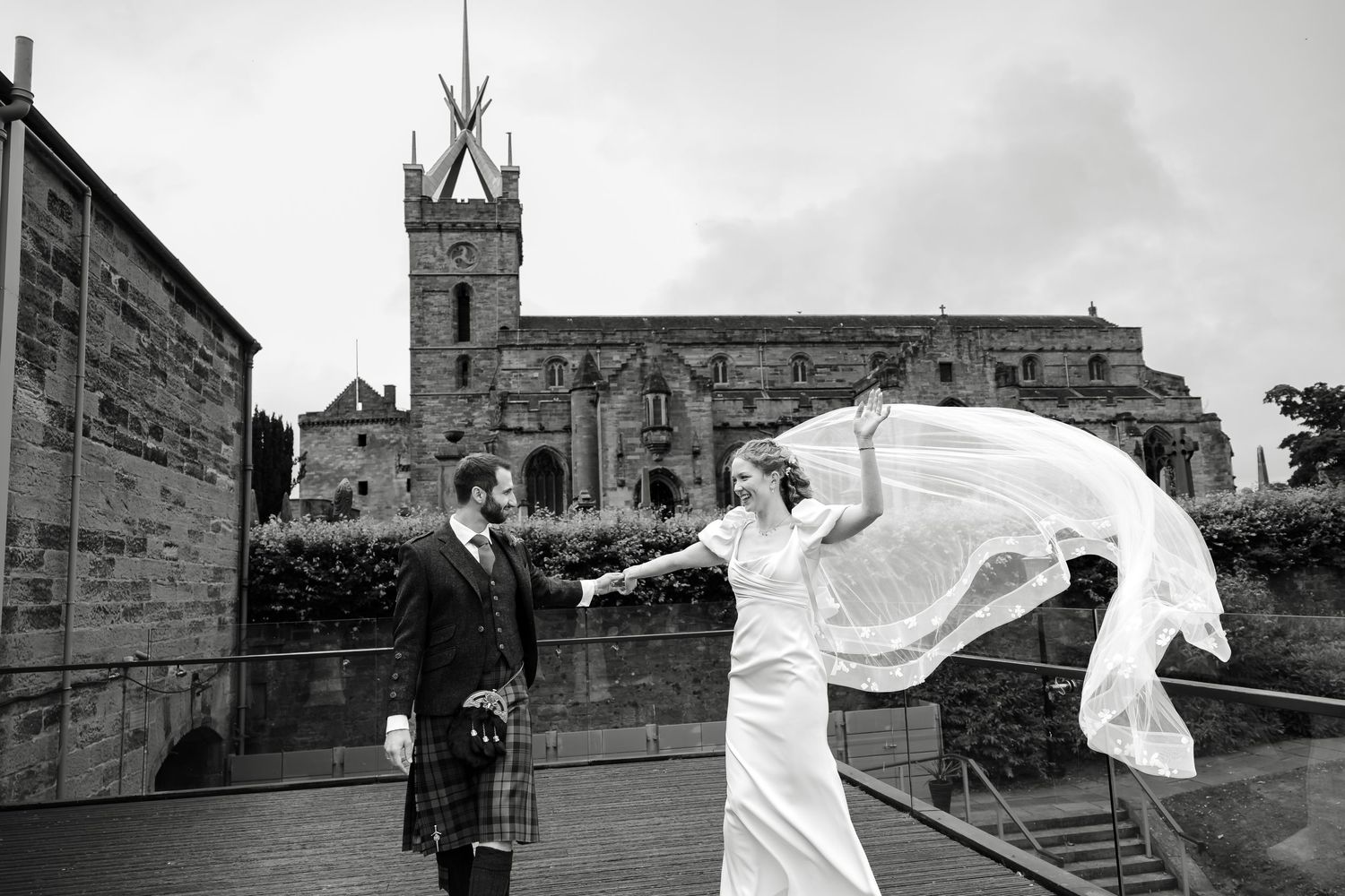 Black and white wedding photo of bride and groom outside historic Scottish church, bride's veil flowing in wind.