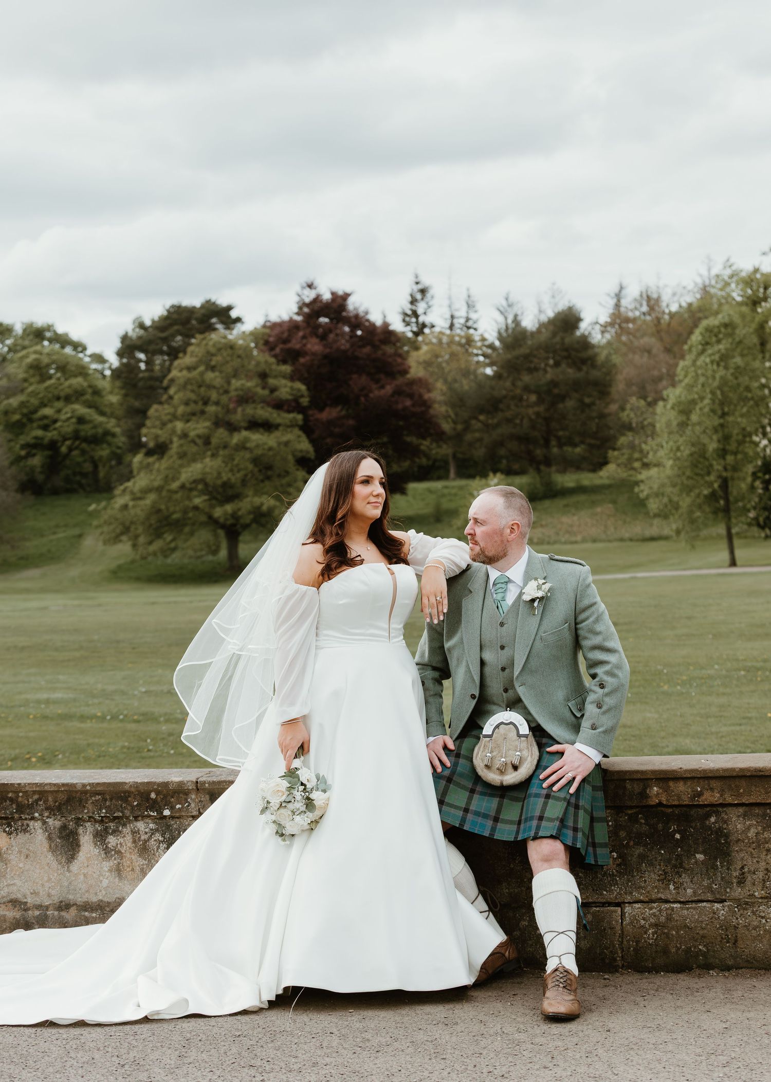 Bride in white gown and groom in green tartan kilt pose together outdoors at a Scottish wedding venue.