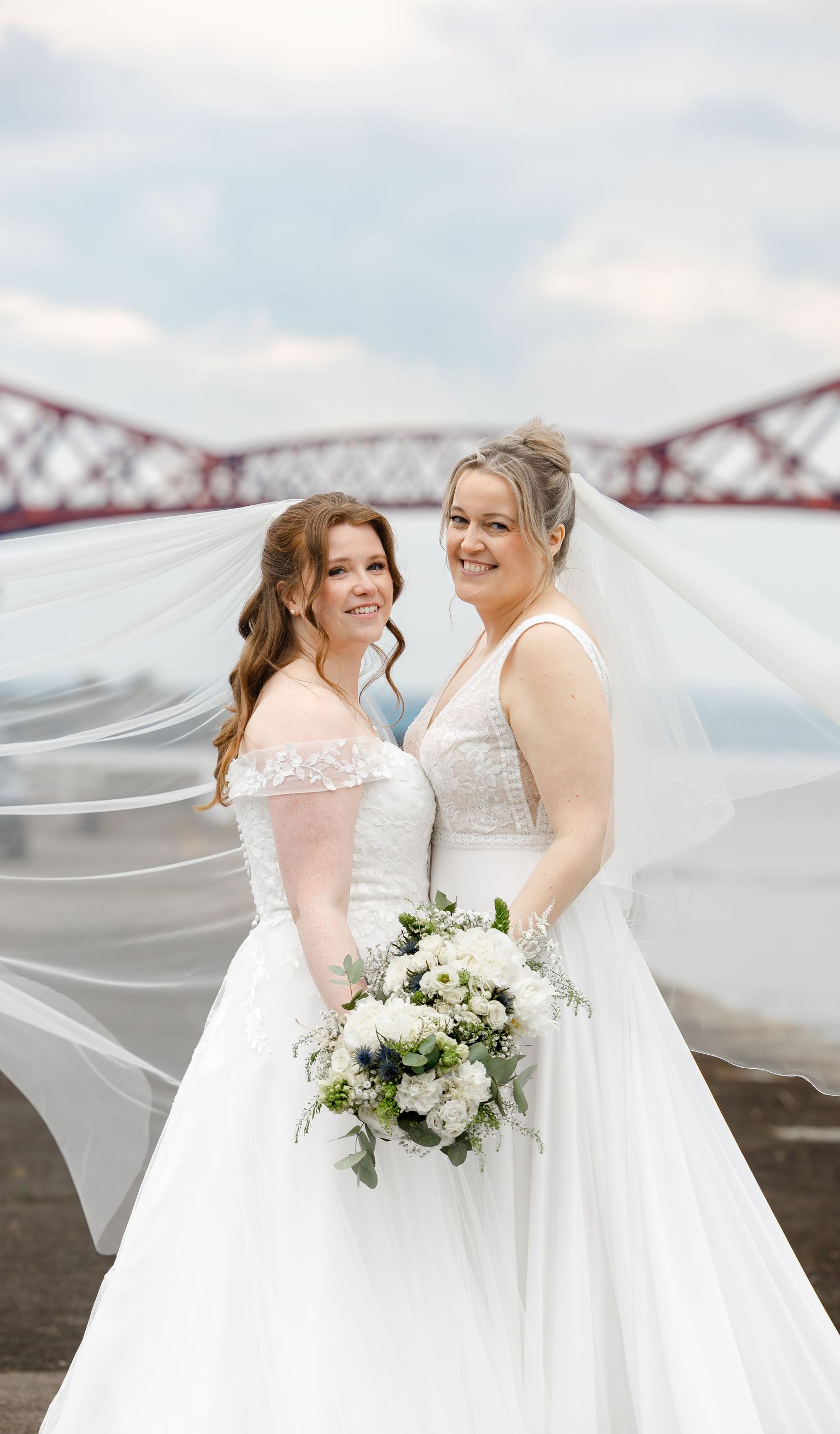 Two brides in white wedding gowns pose with a bouquet near the Forth Bridge, Scotland.