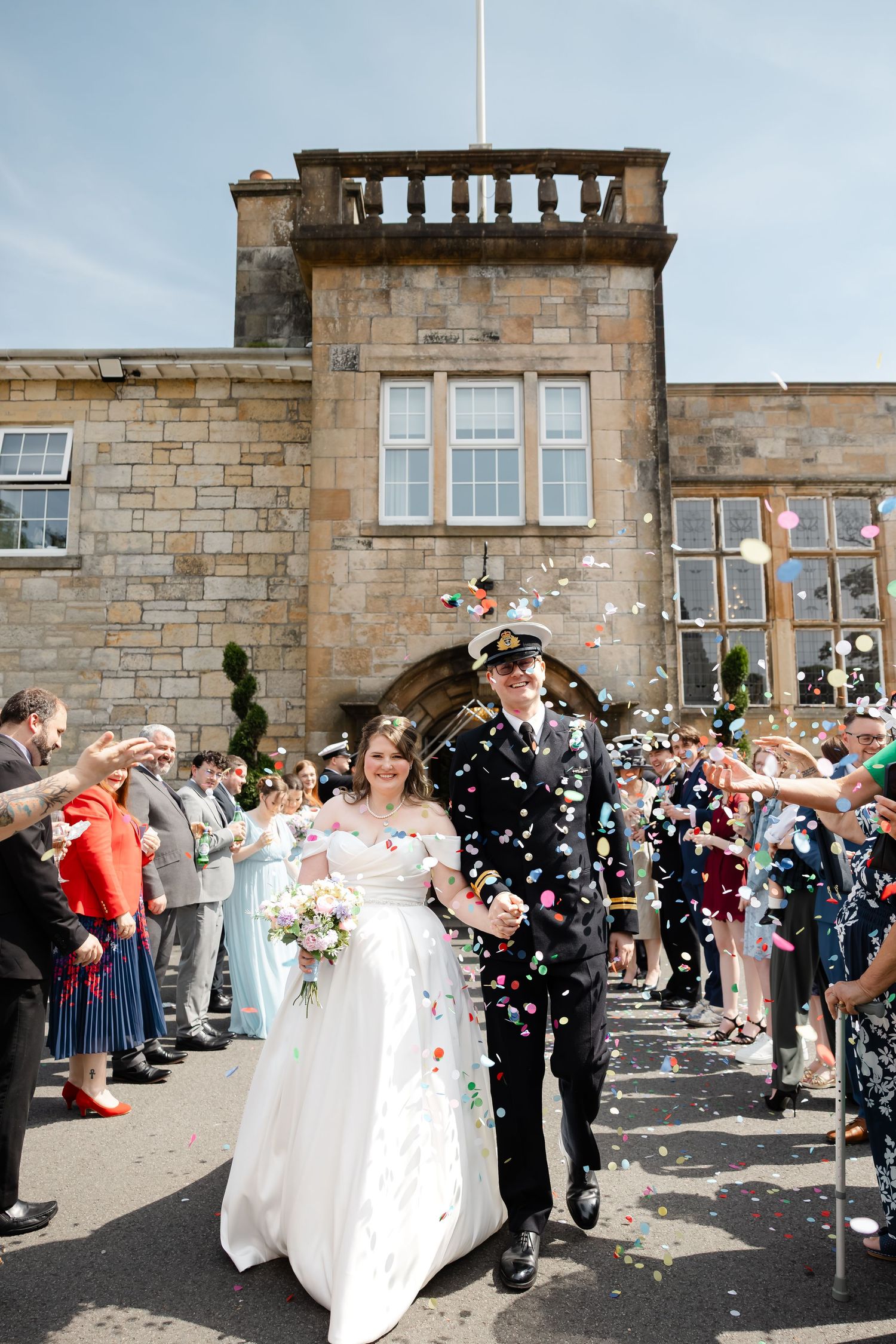 Newlywed couple walking through confetti shower outside a historic stone venue surrounded by wedding guests.
