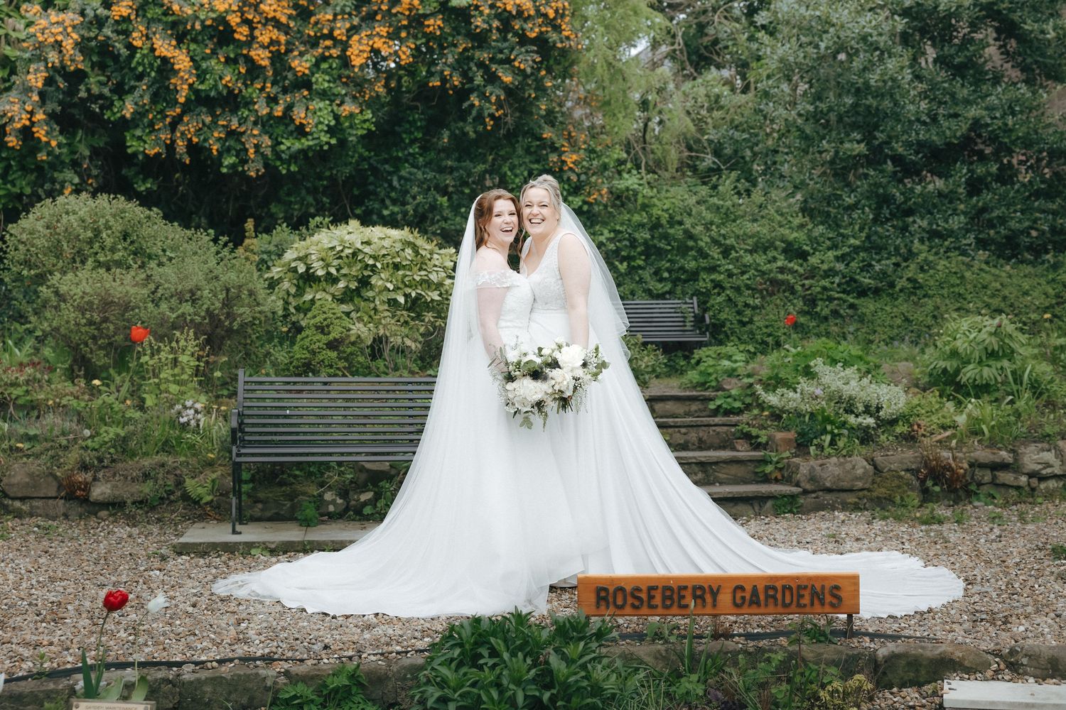 Two brides in white wedding gowns and veils pose together at Roseberry Gardens surrounded by lush greenery.