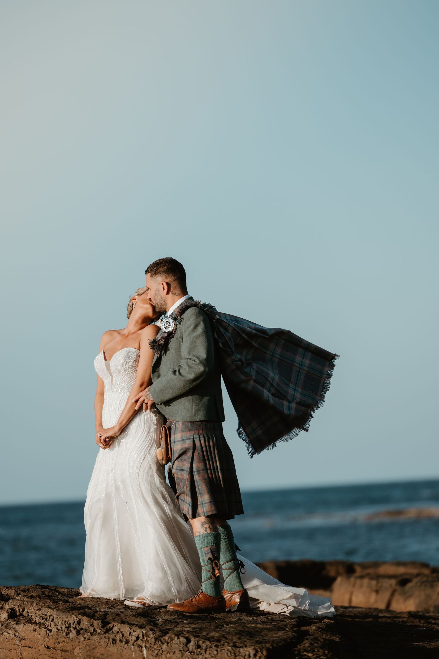 Bride and groom kissing on rocky shore, groom in tartan kilt with sash blowing in wind, ocean backdrop.