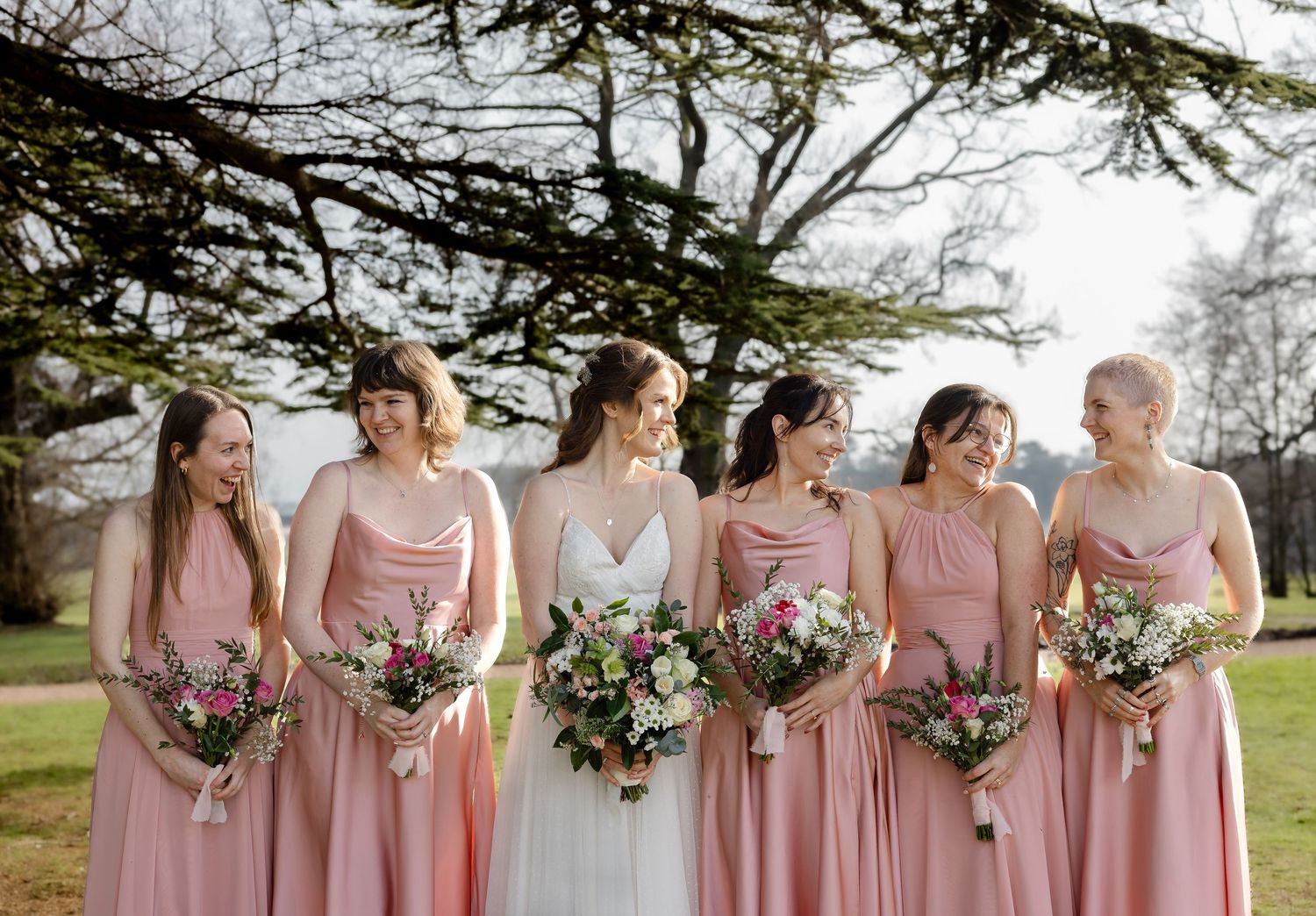 A wedding party in blush pink dresses poses together outdoors while holding floral bouquets.