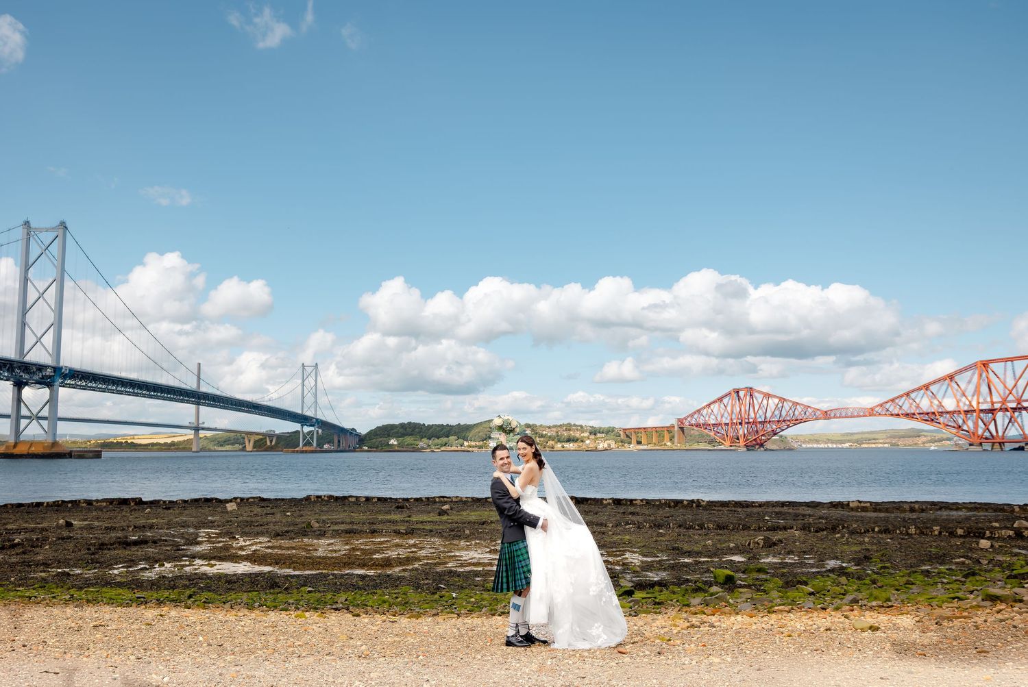 Wedding couple embracing near the Forth Bridge and Forth Road Bridge in Scotland on a sunny day.