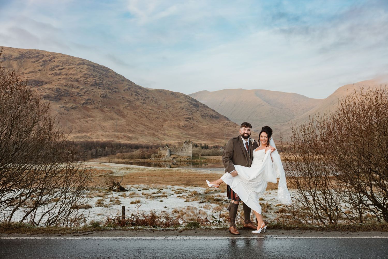 A couple in wedding attire poses dramatically against a backdrop of snow-covered Scottish highlands. Inveraray