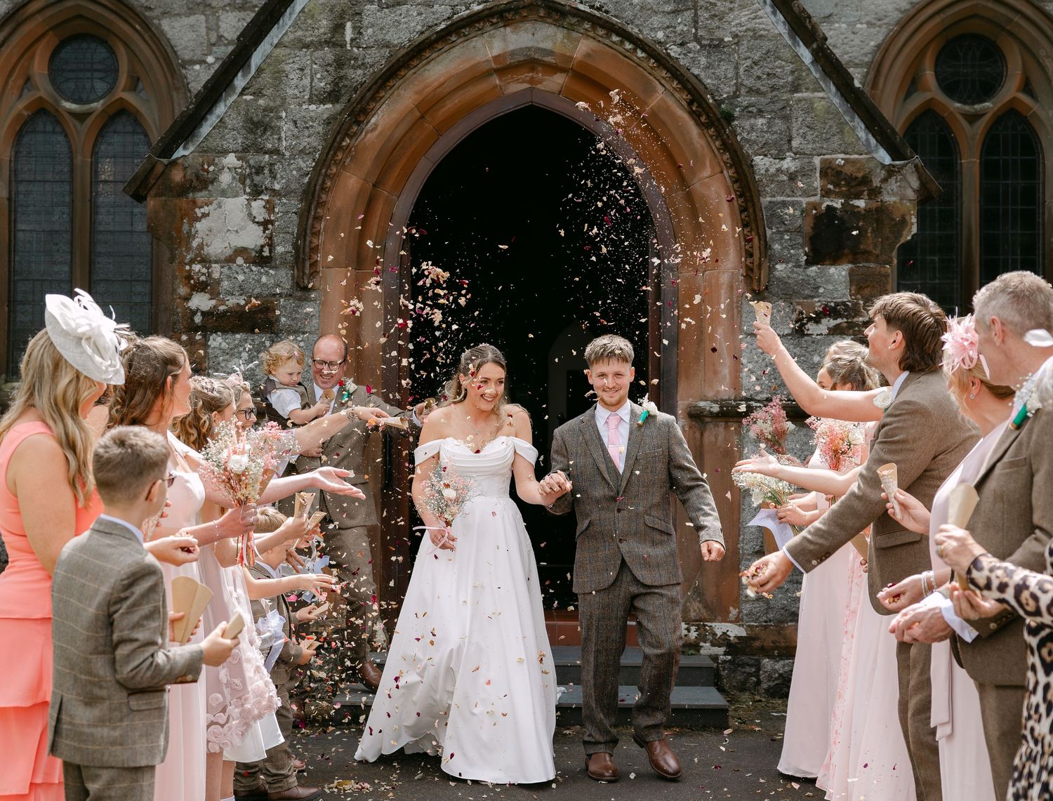 Bride and groom exit church through confetti tunnel surrounded by smiling wedding guests outside stone chapel.