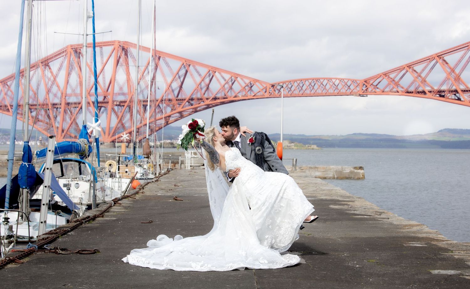 Couple in wedding attire poses romantically on a pier with the iconic Forth Bridge in the background in Scotland.