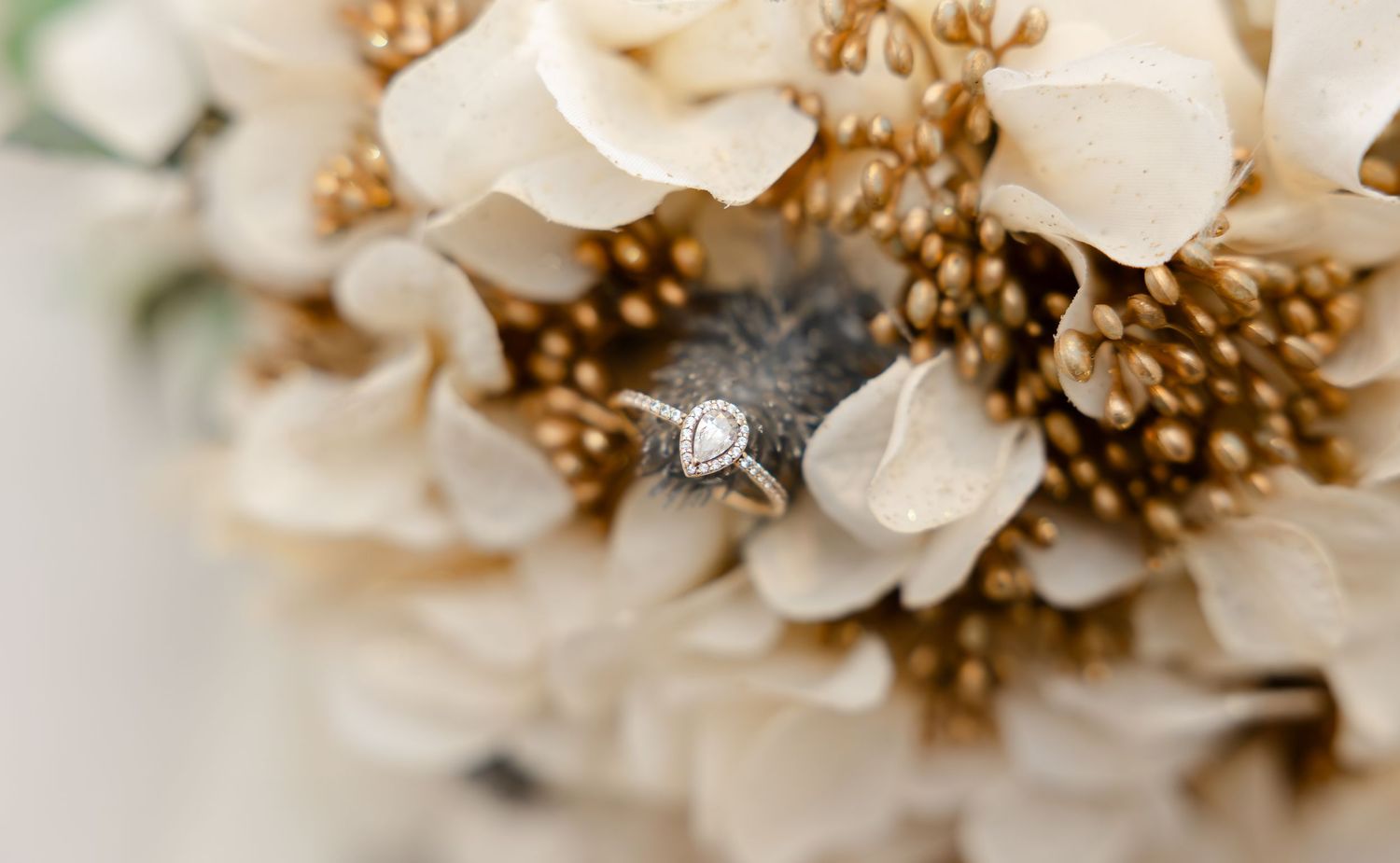 Close-up of a diamond halo engagement ring nestled in a bridal bouquet of white hydrangeas and gold flowers.