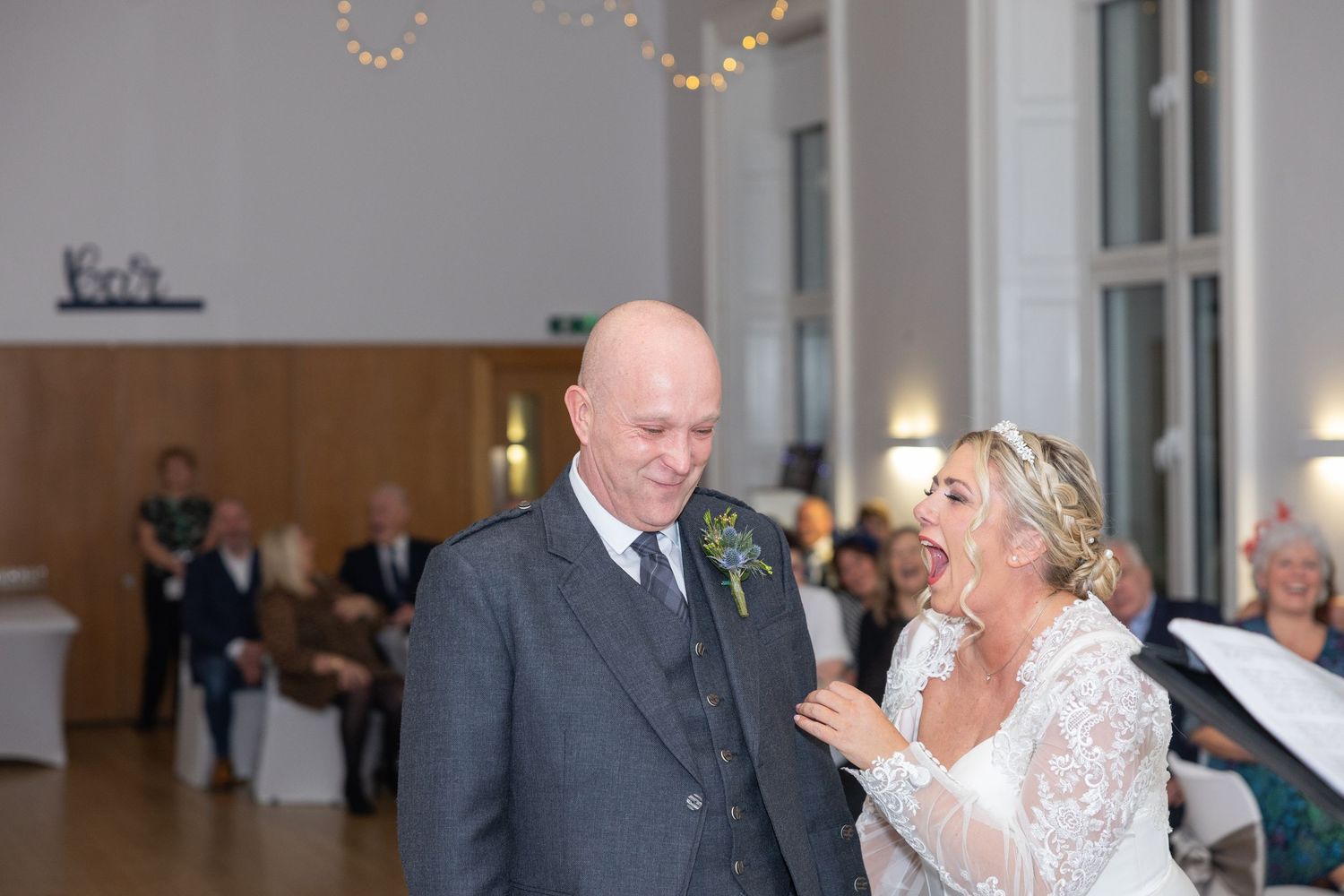 A couple shares a romantic kiss during their wedding ceremony in an elegant white venue with guests looking on.