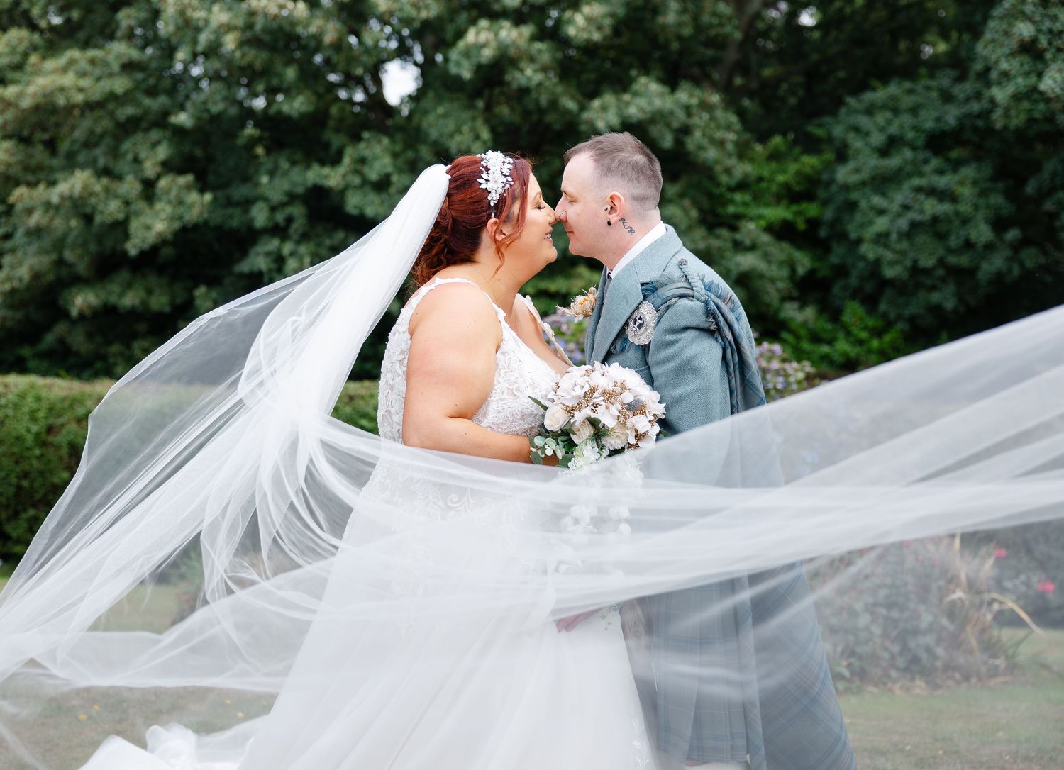 Bride and groom sharing a kiss outdoors with wedding veil flowing in the wind, surrounded by lush green trees.