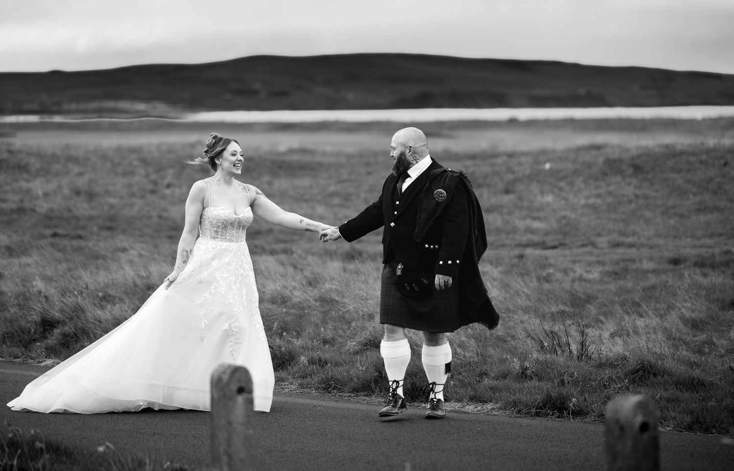 Black and white photo of a bride in white gown and groom in Scottish kilt holding hands outdoors.