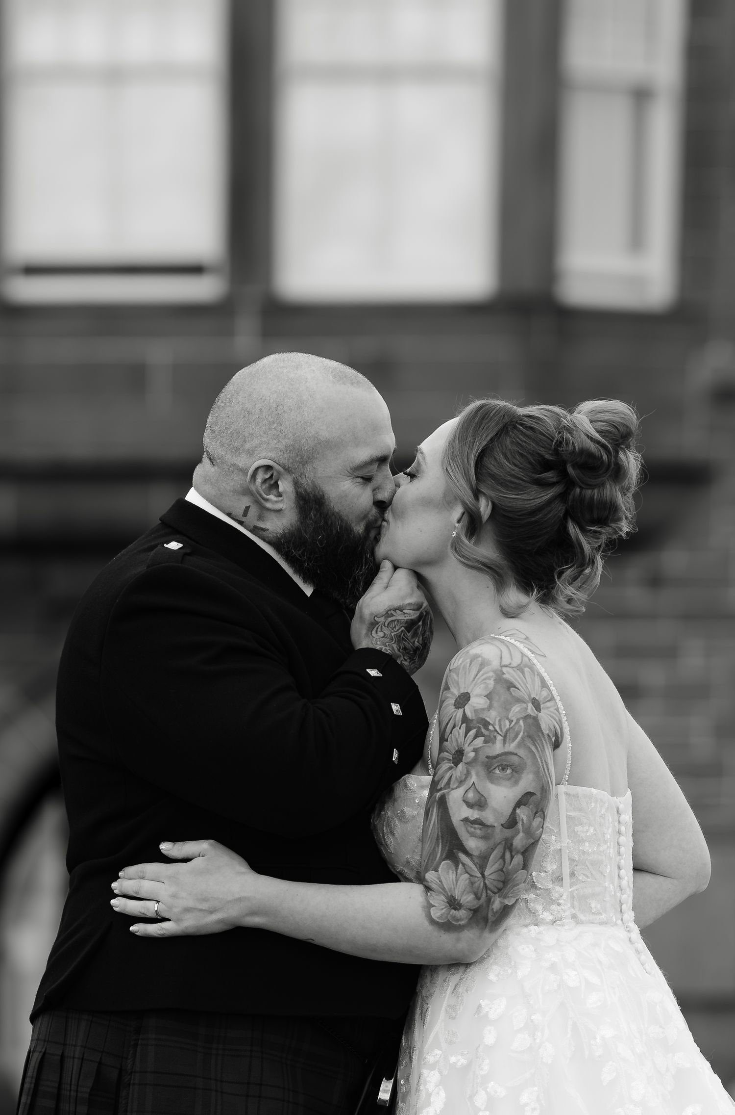Black and white photo of a tattooed bride and groom sharing a kiss in wedding attire outdoors.