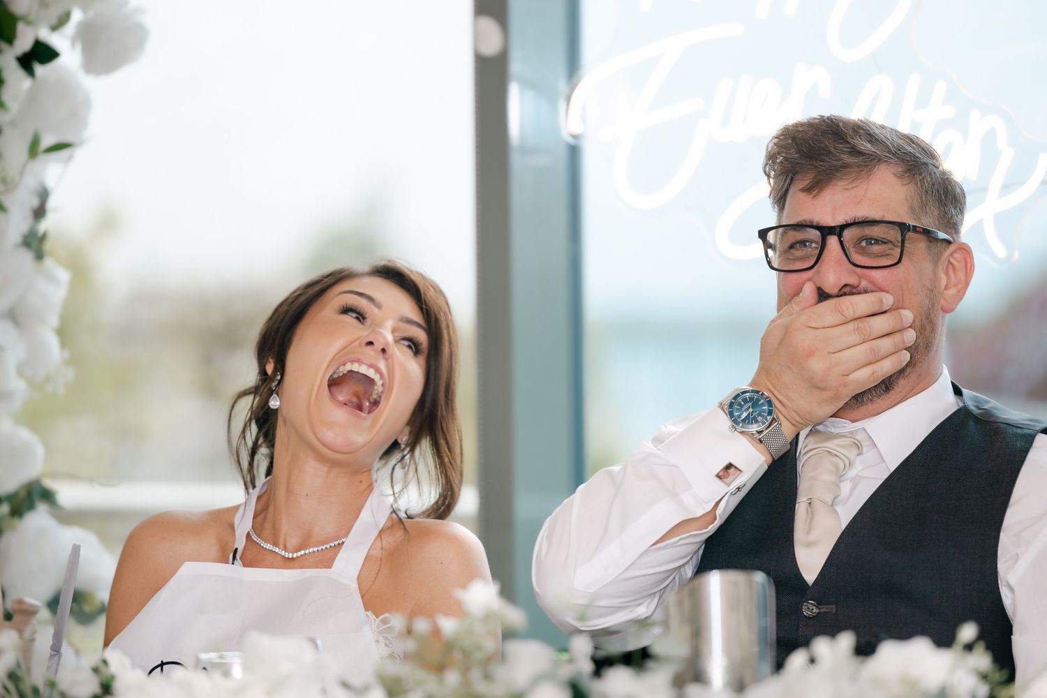 Bride and groom laughing joyfully at wedding reception table adorned with white flowers.