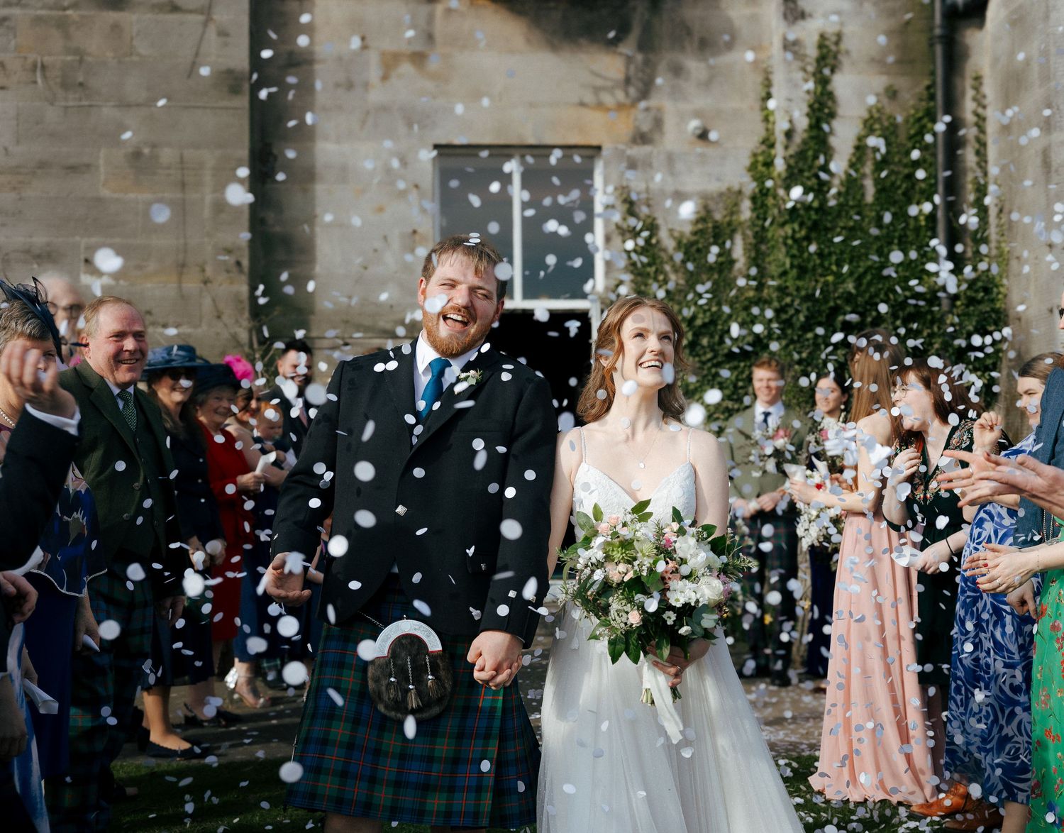 Bride and groom walk through confetti shower outside stone venue, groom in blue tartan kilt, bride holding white floral bouquet.