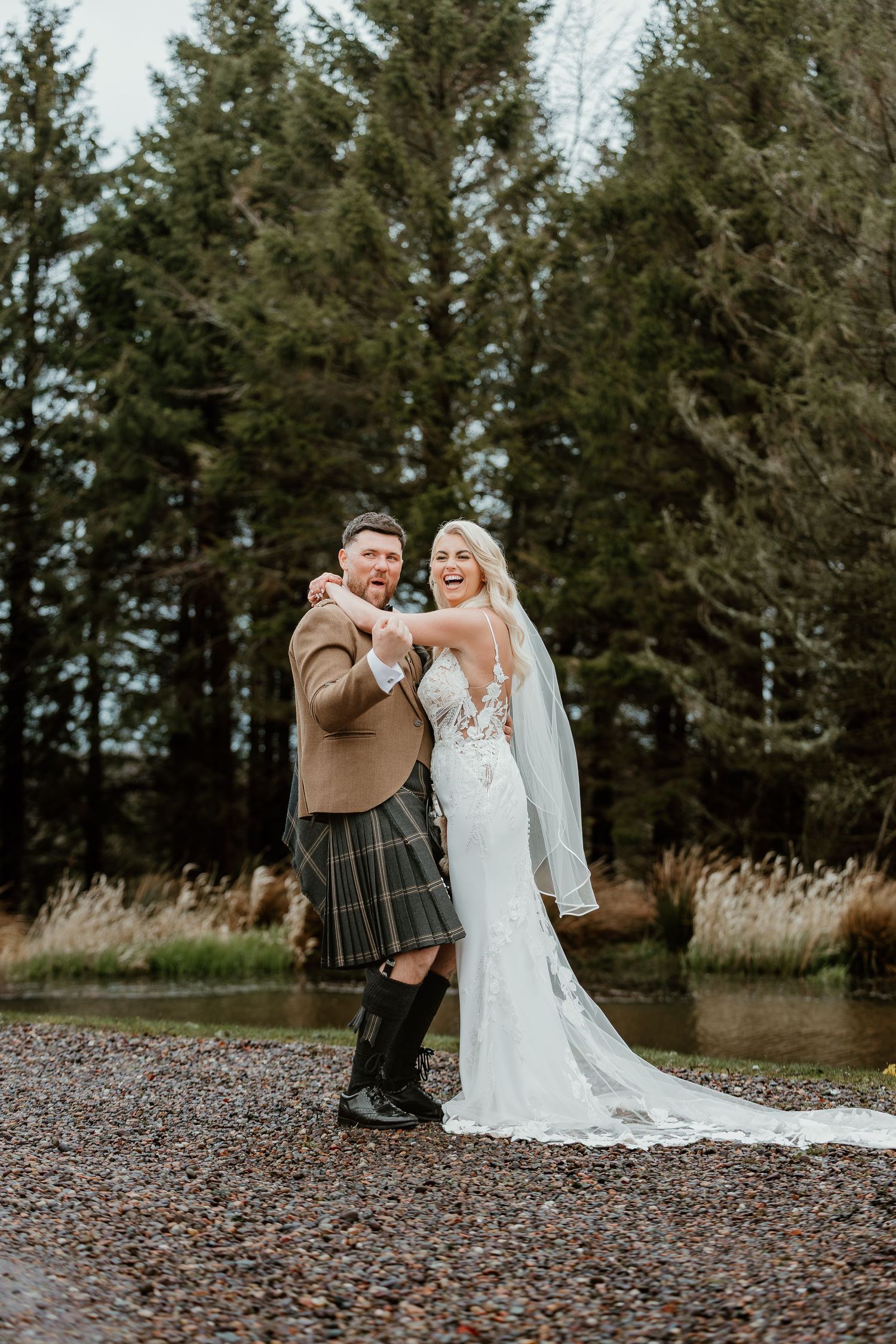 Joyful bride in white lace gown and groom in traditional Scottish kilt dancing outdoors near pine trees.
