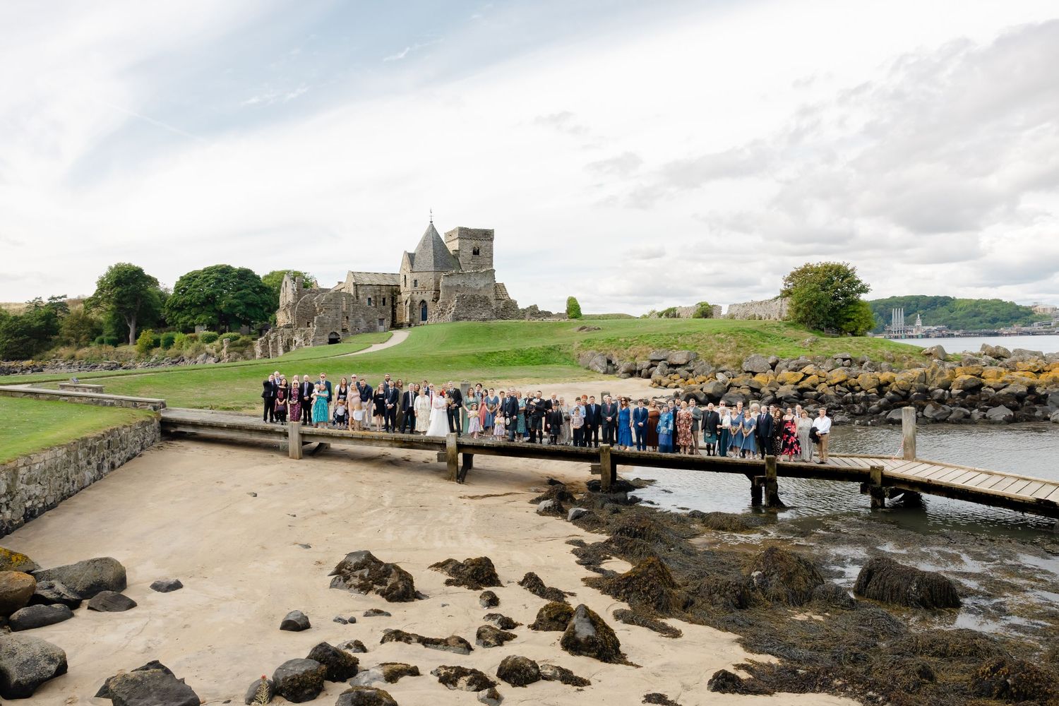 Wedding party gathered on a wooden bridge near Inchcolm Abbey ruins beside the Firth of Forth in Scotland.