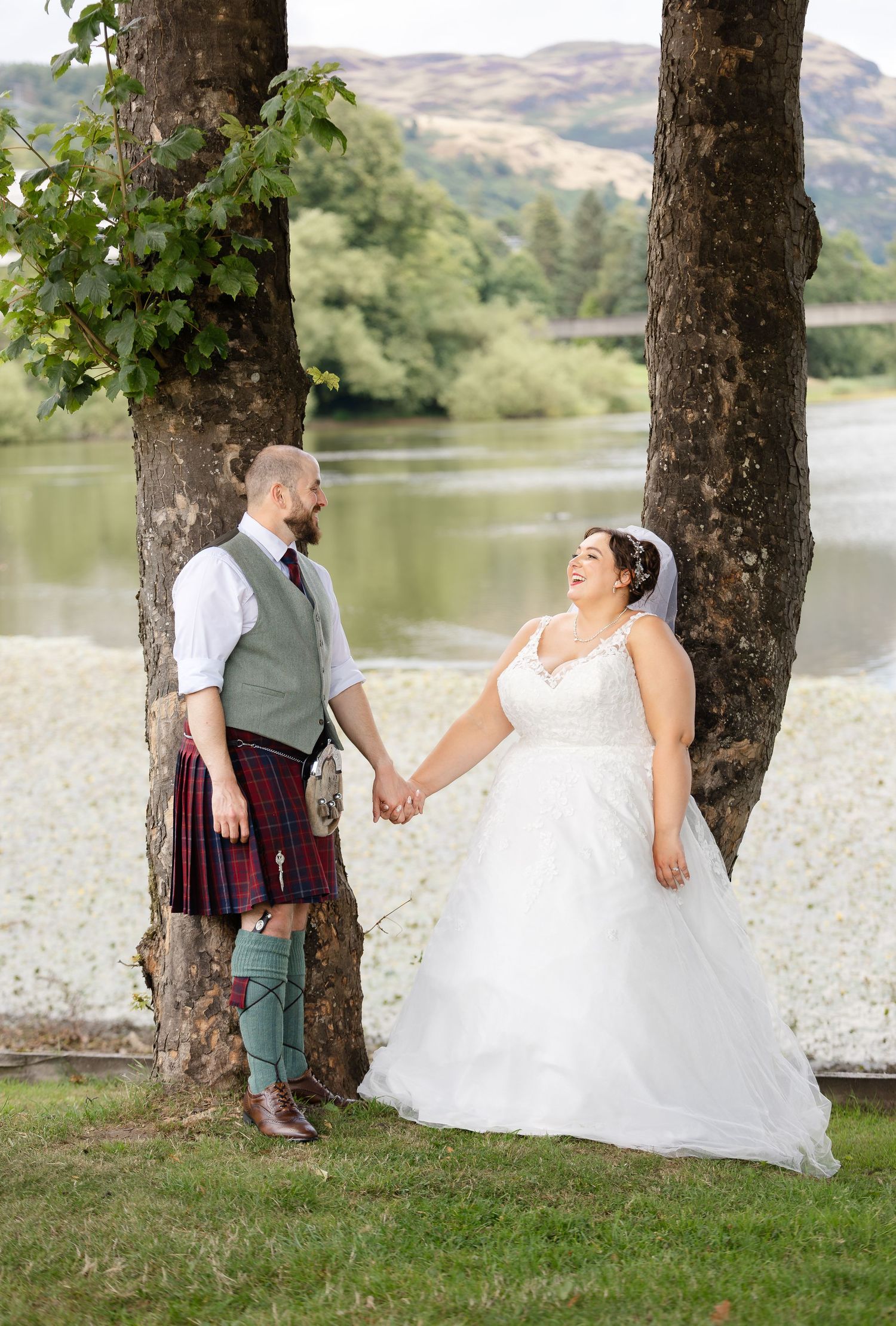 Bride and groom holding hands by a loch in Scotland, groom wearing a red tartan kilt, bride in a white ballgown.