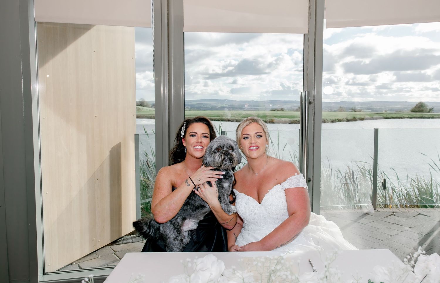 Two brides smile holding a gray dog indoors with scenic waterfront views through large glass windows.