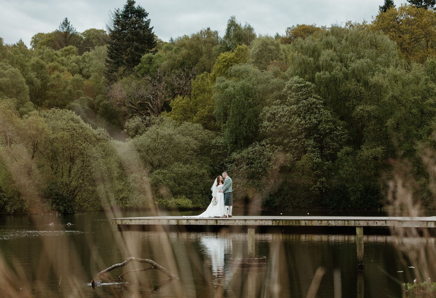 Bride and groom stand on a wooden dock surrounded by a serene lake and lush green trees on their wedding day.