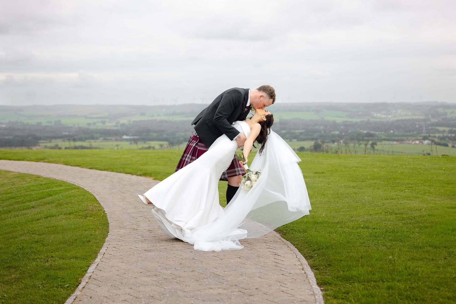 Bride and groom share a romantic dip kiss on a curved garden path with lush green hills in the background.