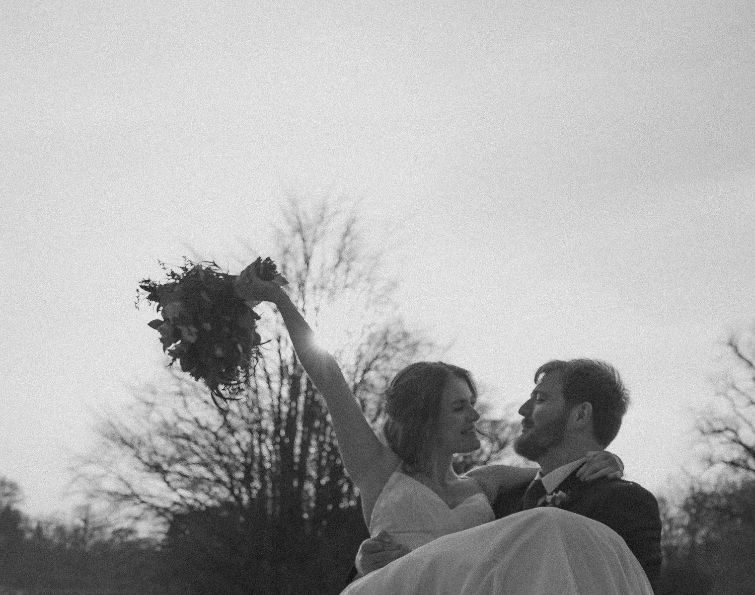 Black and white wedding photo of groom carrying bride who raises bouquet joyfully outdoors among bare trees.